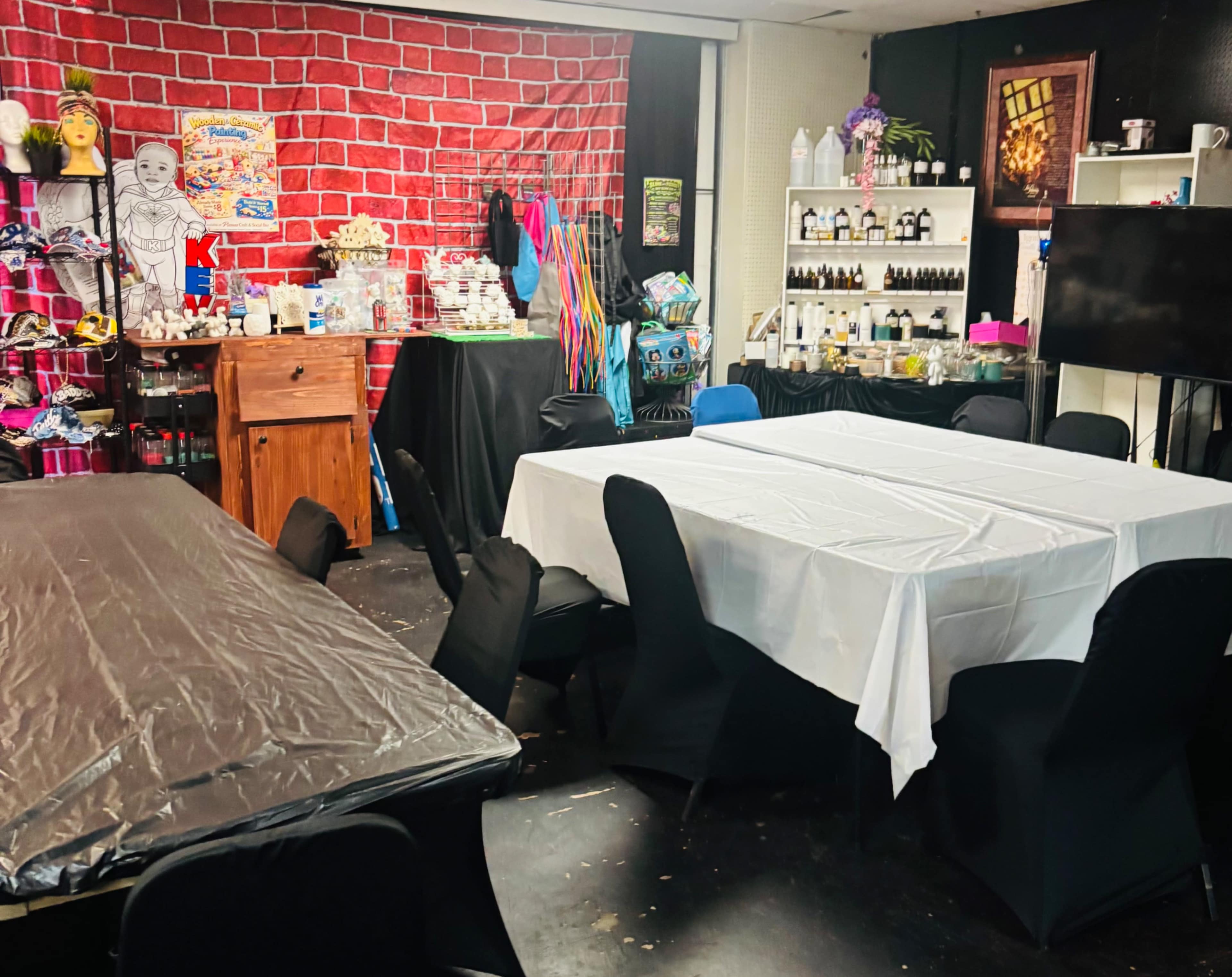 The image shows a room set up with multiple tables covered in white and black tablecloths, against a backdrop of a red brick wall and various decorative items on shelves.