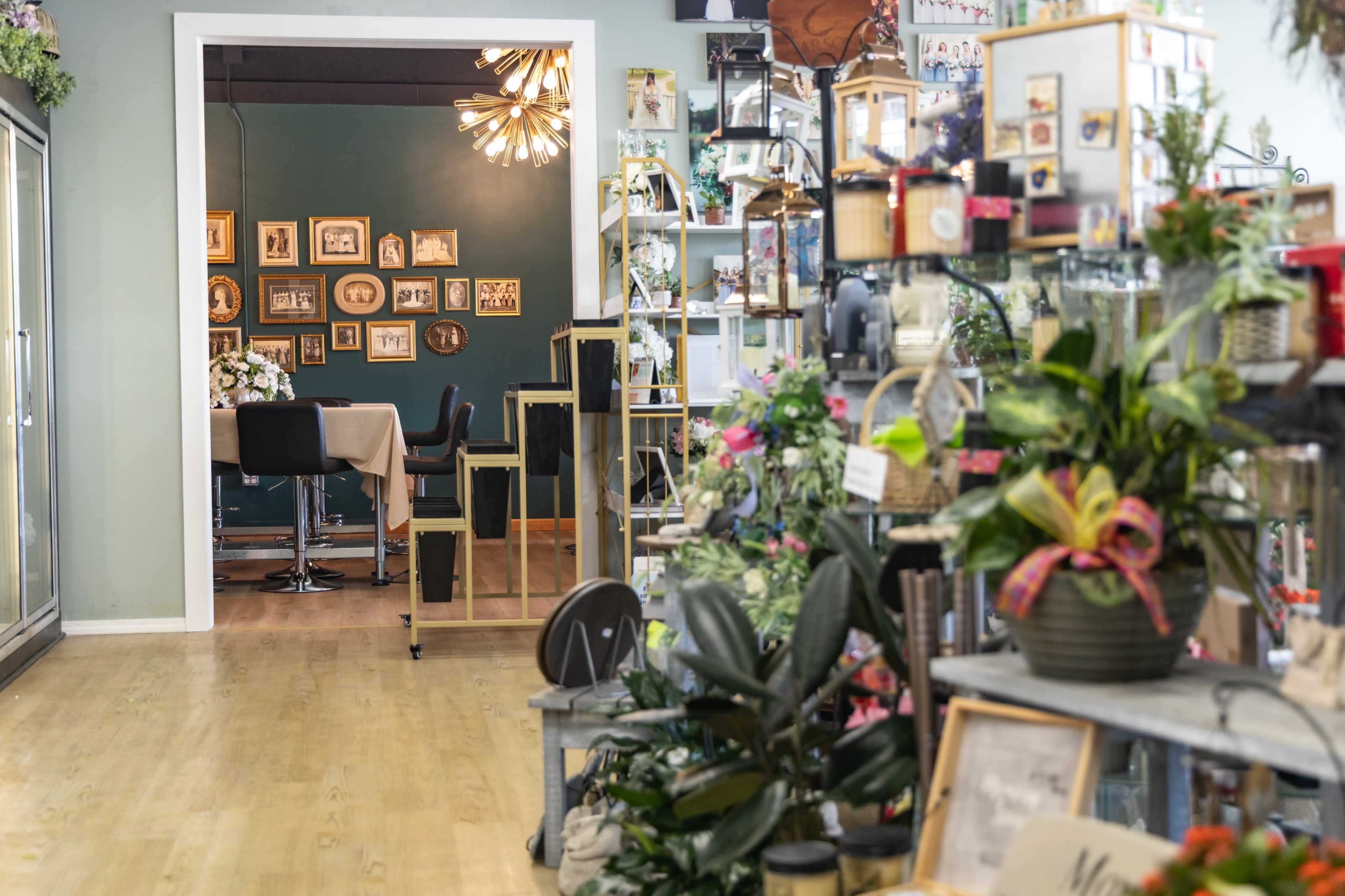The image shows a bright interior space featuring a pathway lined with potted plants and decorative items, leading to a room with framed pictures on the wall and a dining table.