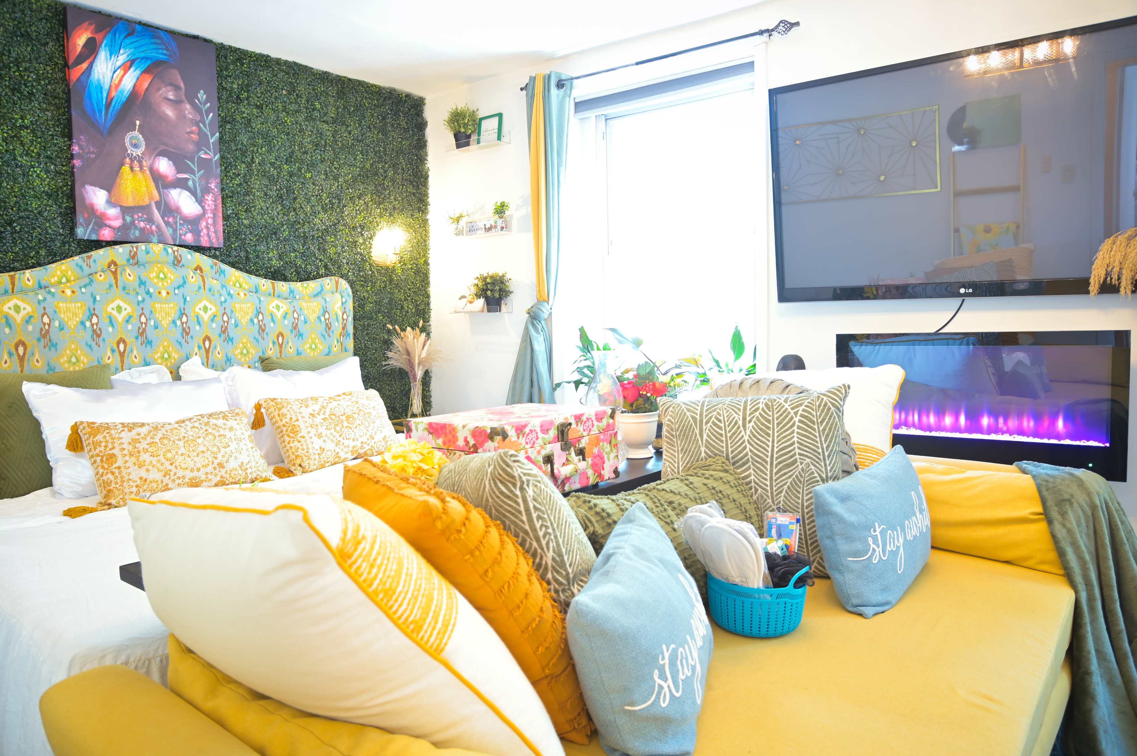 The image shows a cozy living space with a patterned yellow and white sofa, decorated with various cushions and plants, and a wall-mounted television above a modern electric fireplace.
