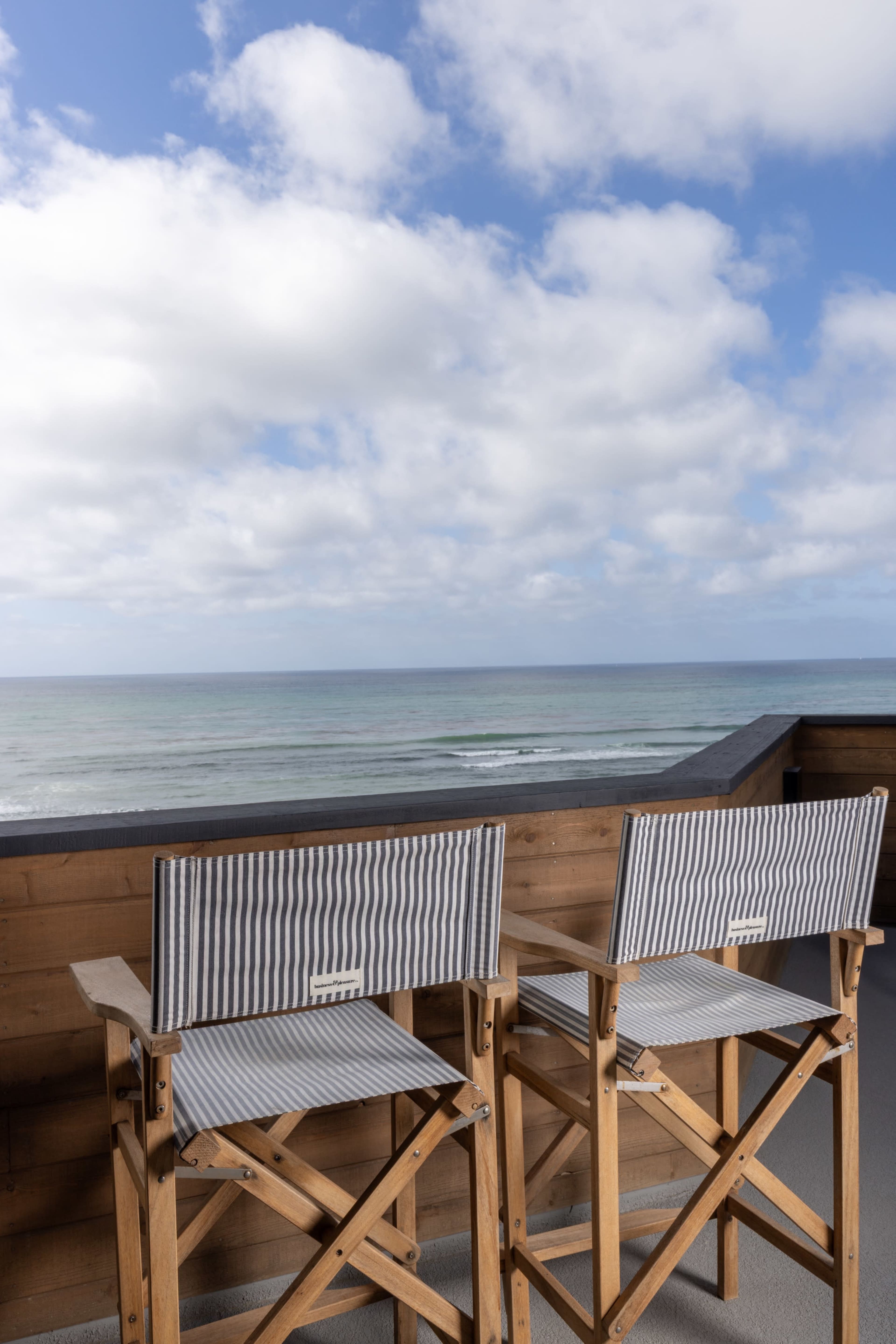Two wooden deck chairs with striped fabric are positioned on a balcony overlooking the ocean under a partly cloudy sky.