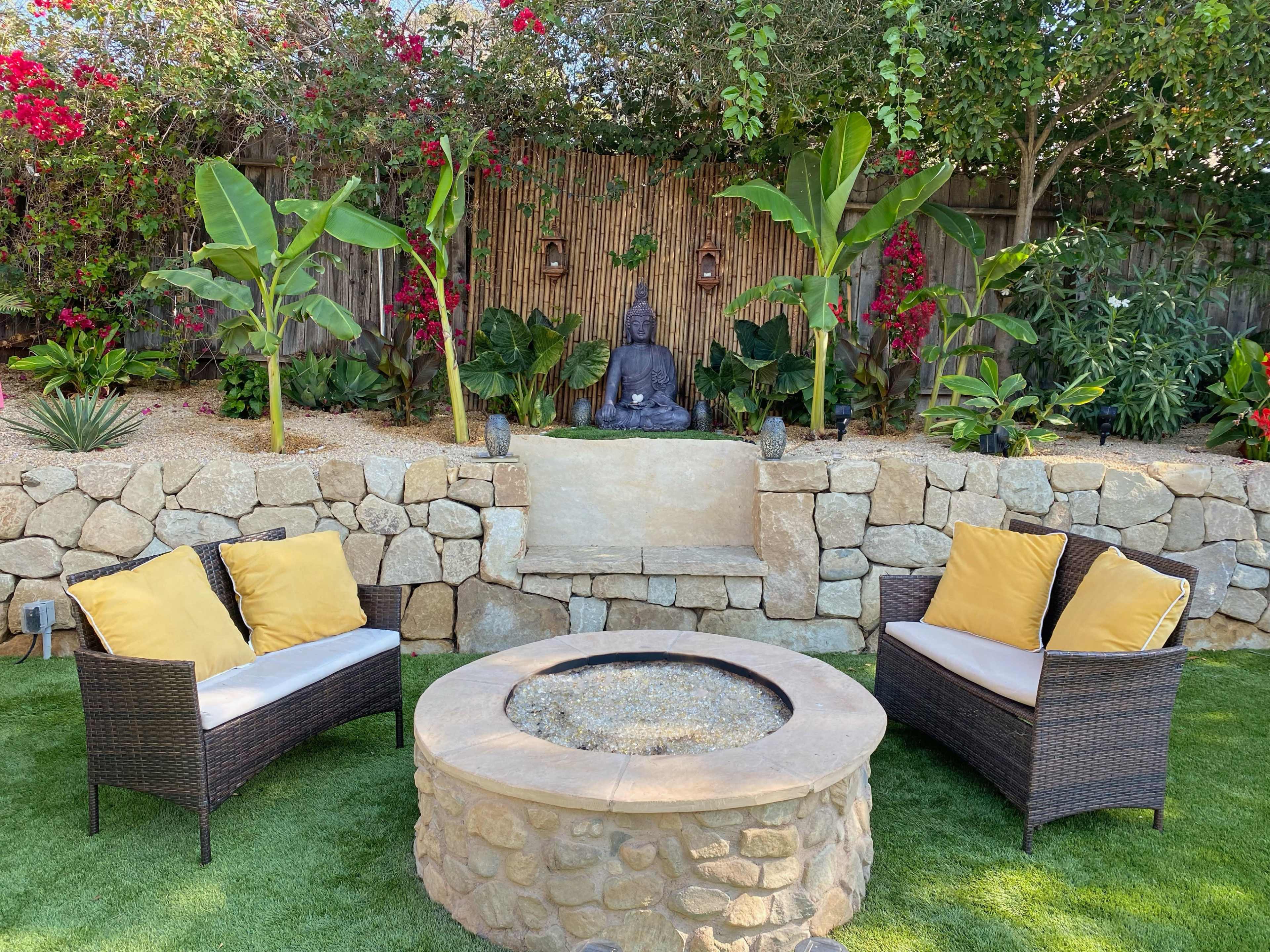 A cozy outdoor seating area features two wicker chairs with yellow cushions, facing a stone fire pit and a decorative Buddha statue surrounded by lush plants and vibrant flowers.