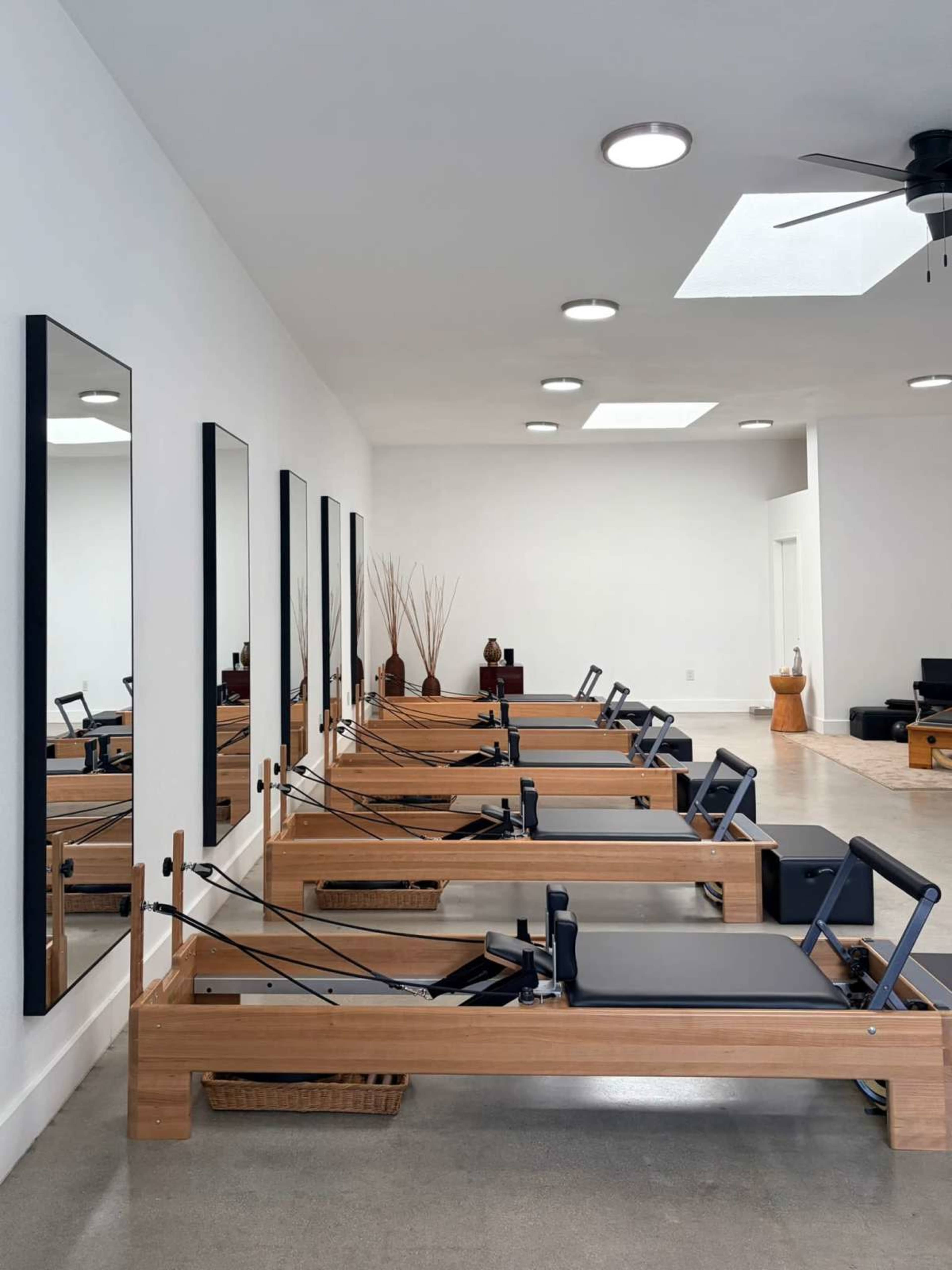 The image shows a reformer gym studio with several wooden Pilates reformers aligned in rows facing large mirrors on a white wall.