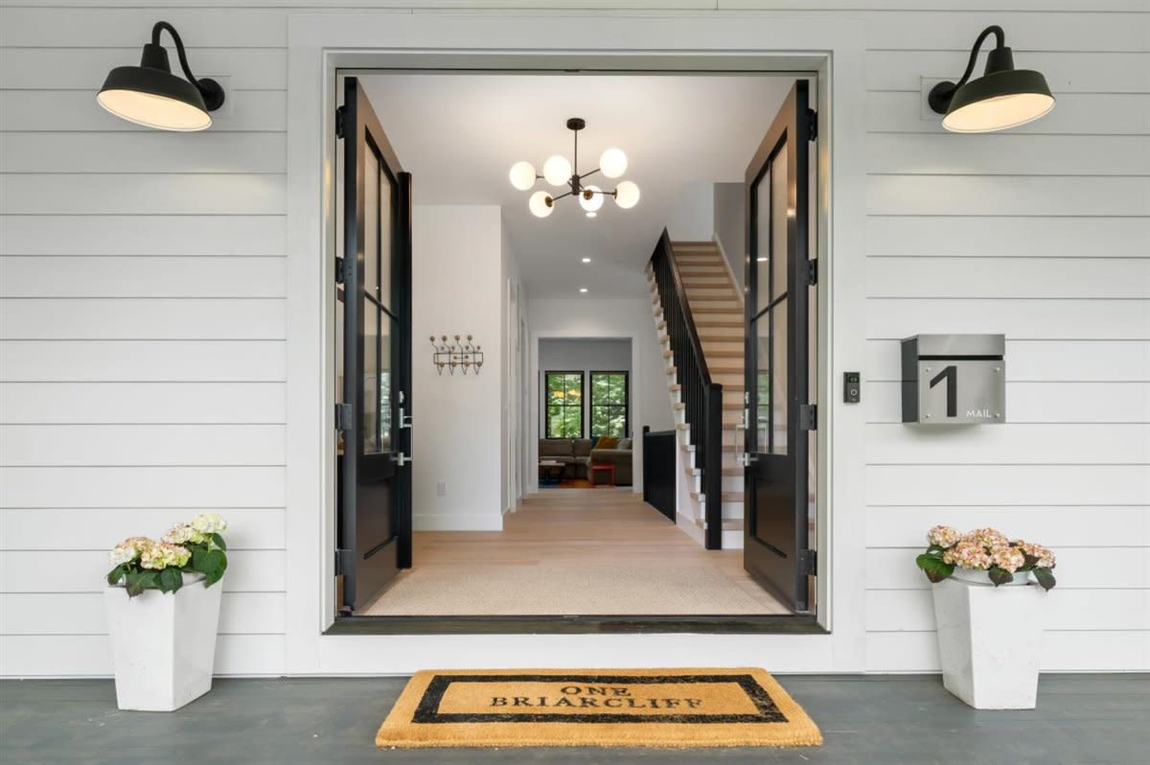 The entrance of a modern home with a clearly marked doorway, a welcome mat, and two light fixtures, leading into an open foyer with stairs visible in the background.