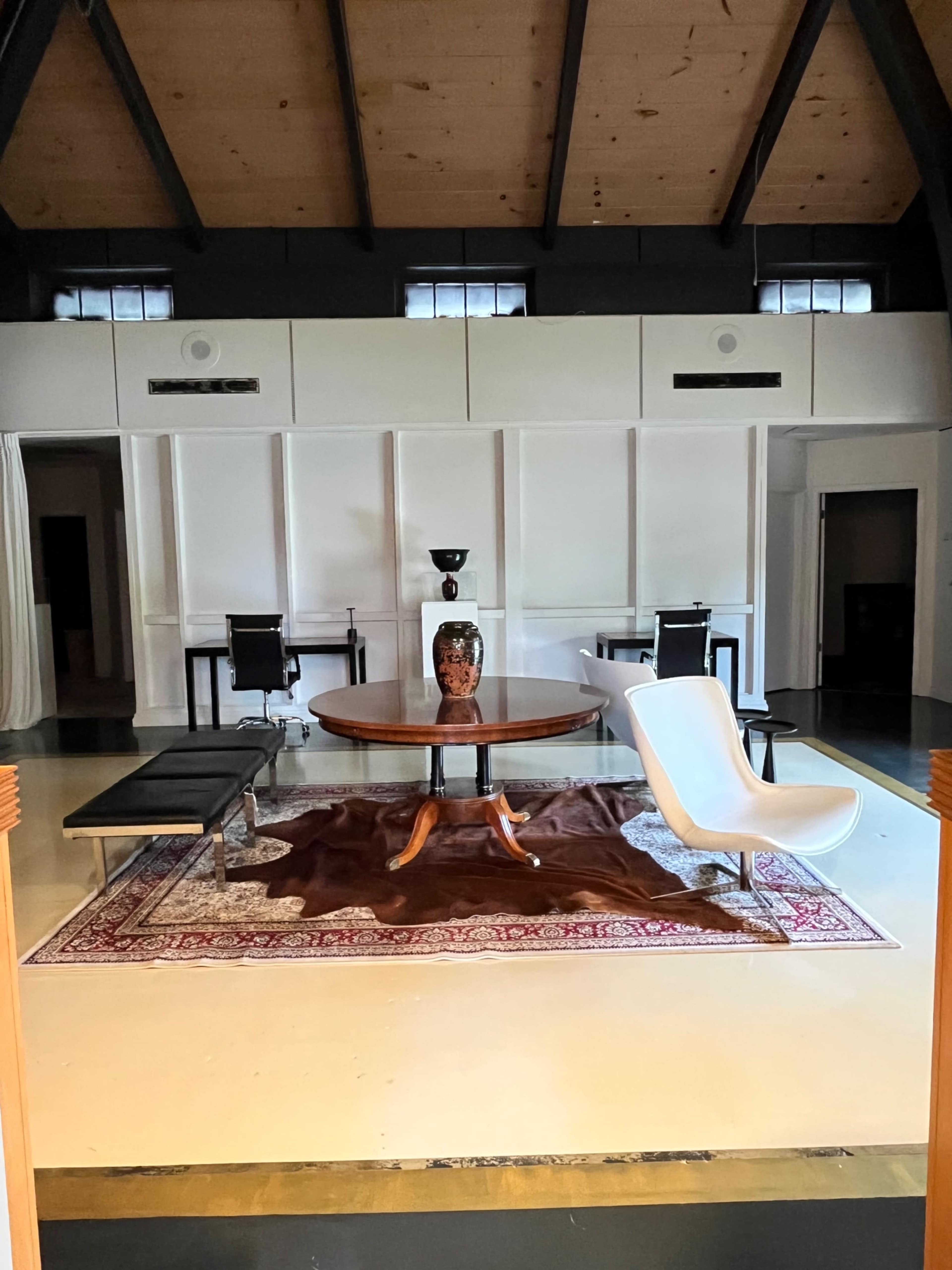 The image shows a spacious room with a wooden ceiling, a round table in the center, and modern furniture arranged on a patterned rug.