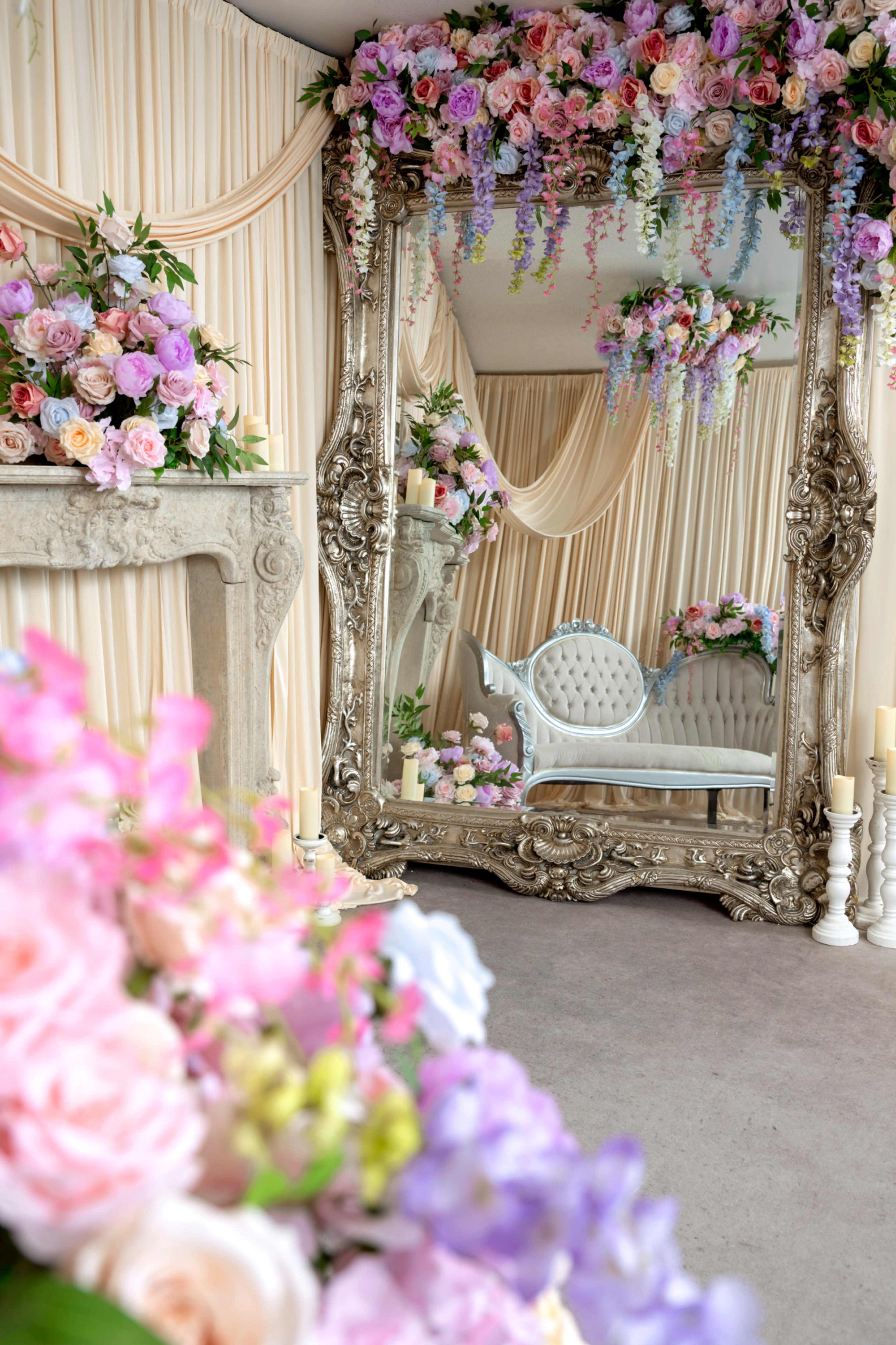 The image features an elegantly decorated interior with a large ornate mirror reflecting a vintage-style couch, surrounded by floral arrangements and draped fabric.