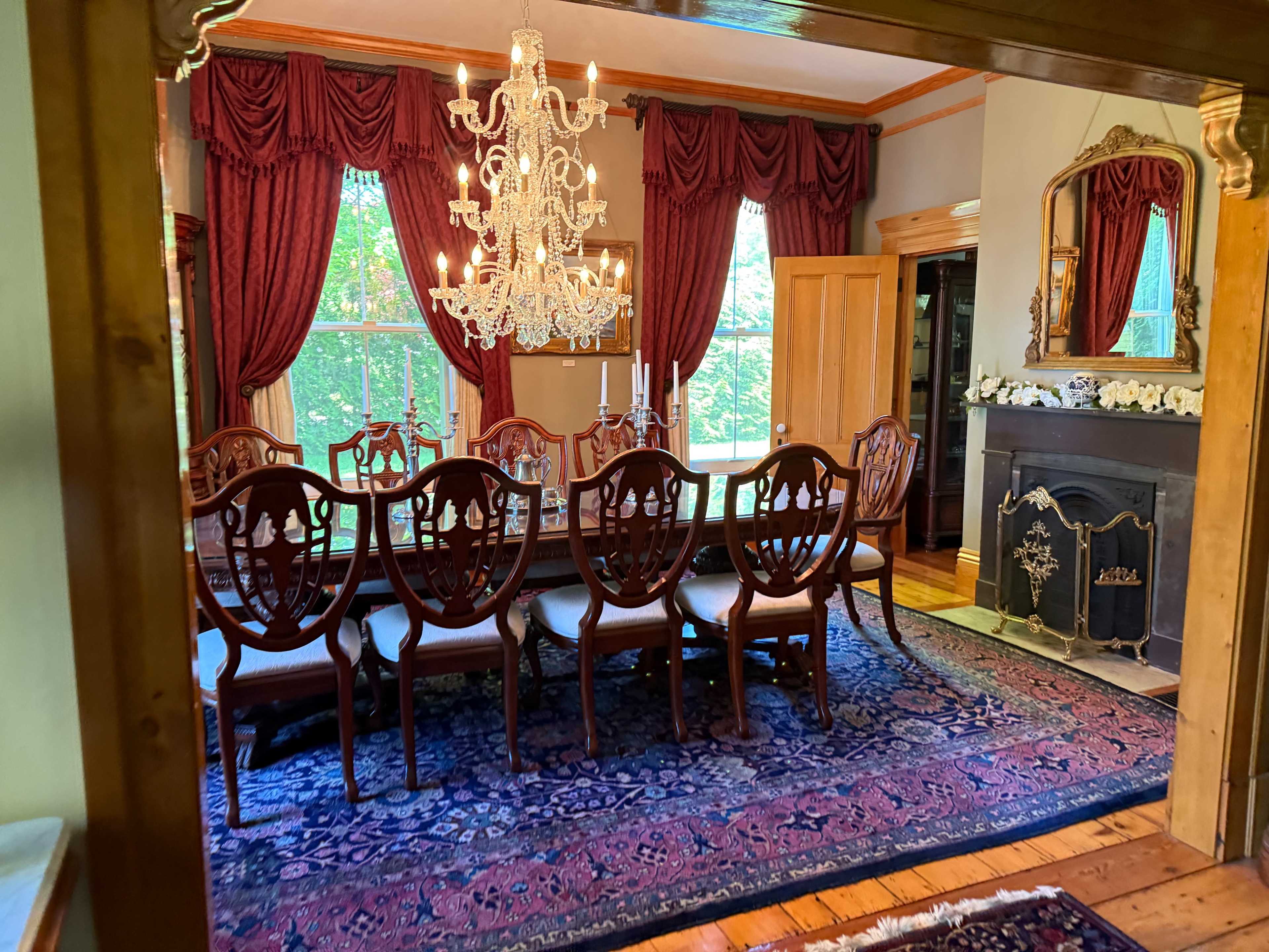 A spacious dining room features a long wooden table surrounded by ornate chairs, a chandelier hangs above, and large windows are draped with rich curtains.