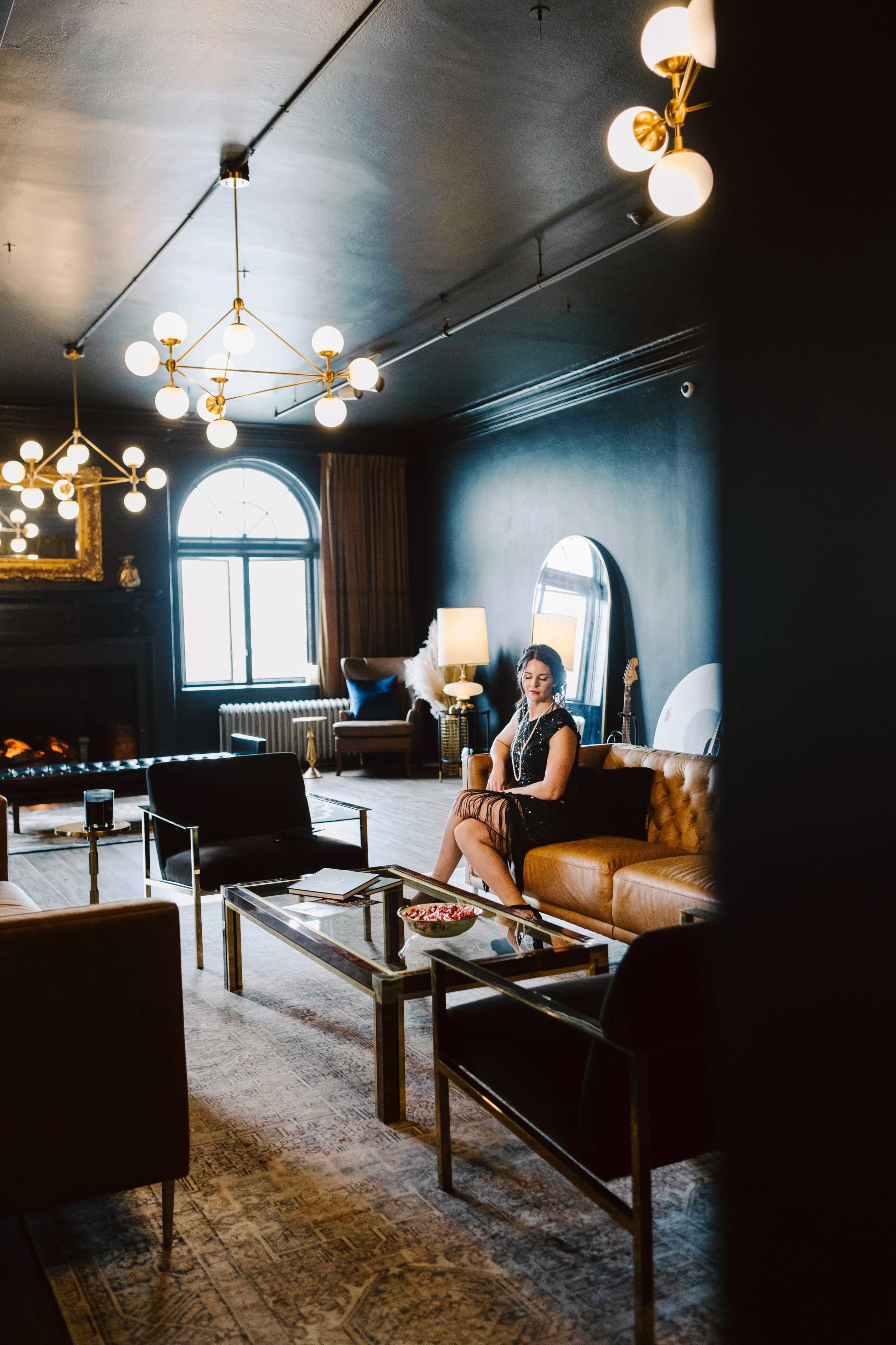 A woman sits on a brown leather sofa in a dark, stylish lounge with modern lighting and a large window.