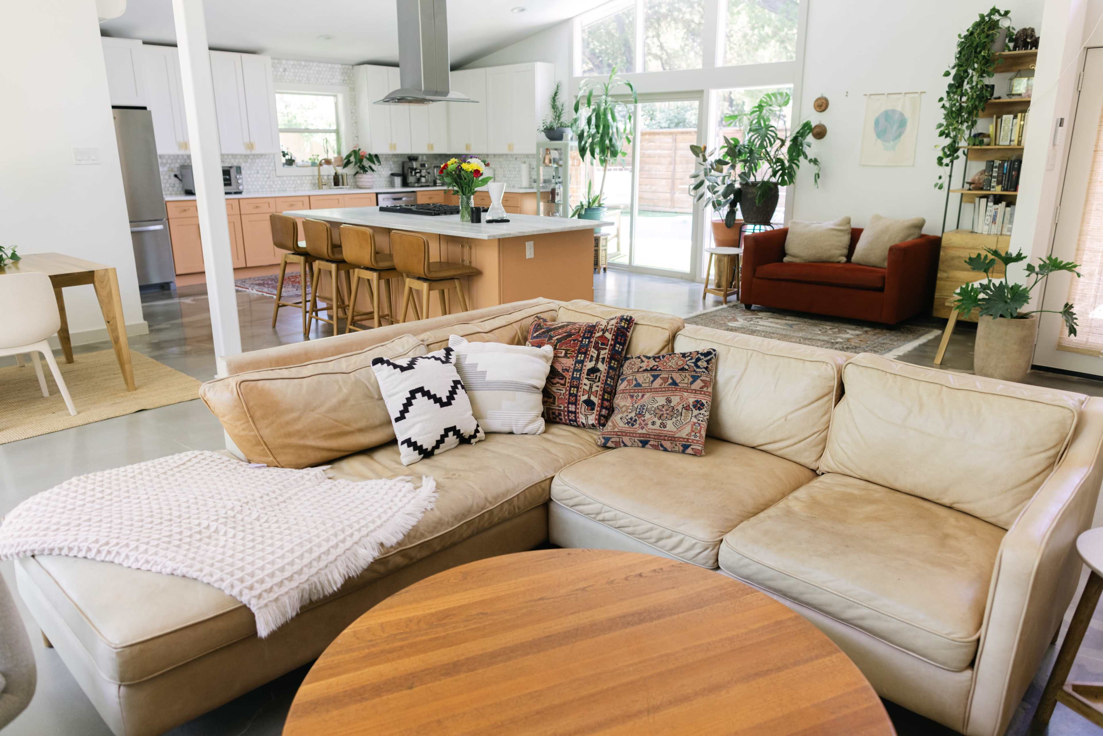 A modern living space featuring a beige sectional sofa, a round wooden coffee table, and an open kitchen with large windows, filled with plants and natural light.