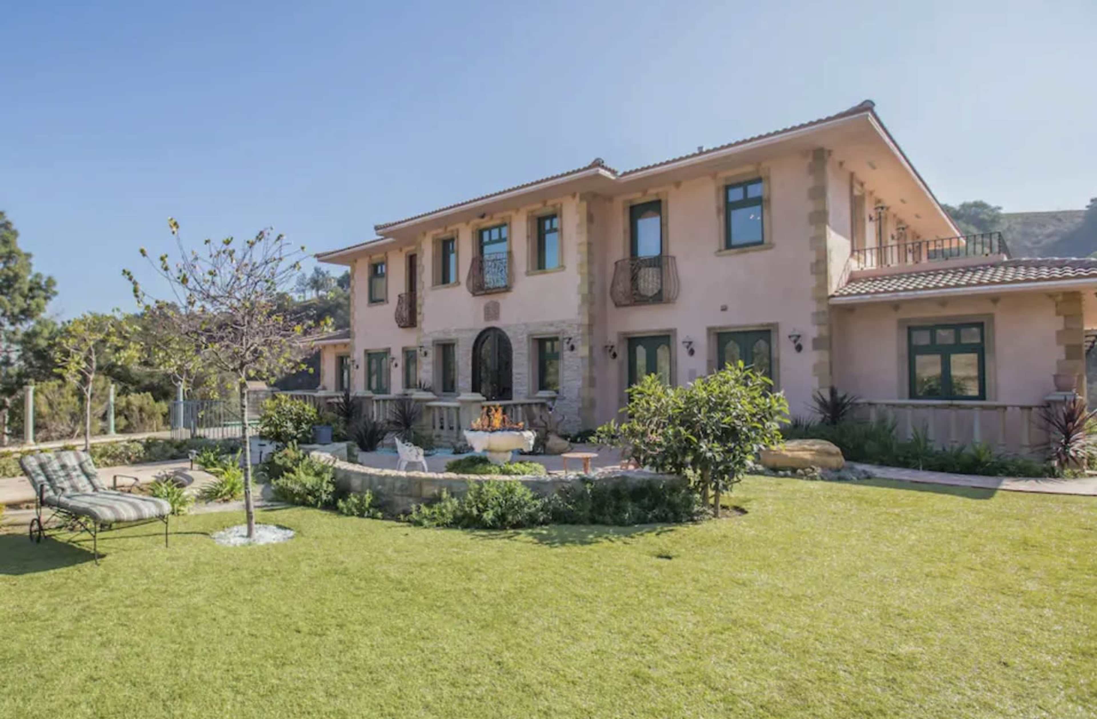 A large two-story house with a landscaped yard, featuring a fountain and a patio area surrounded by greenery.