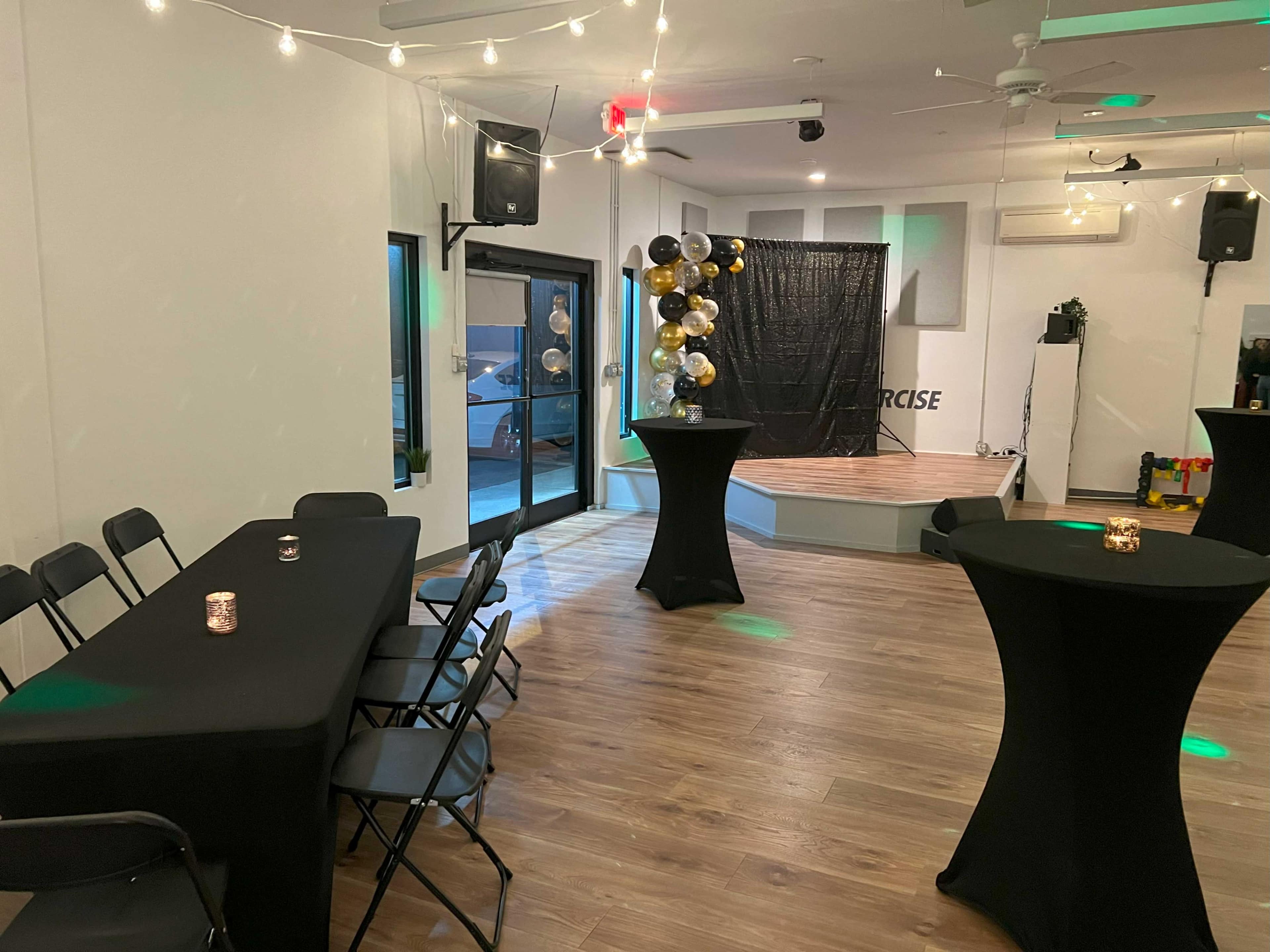 An empty event space with black tables and a stage decorated with black and gold balloons in the background.