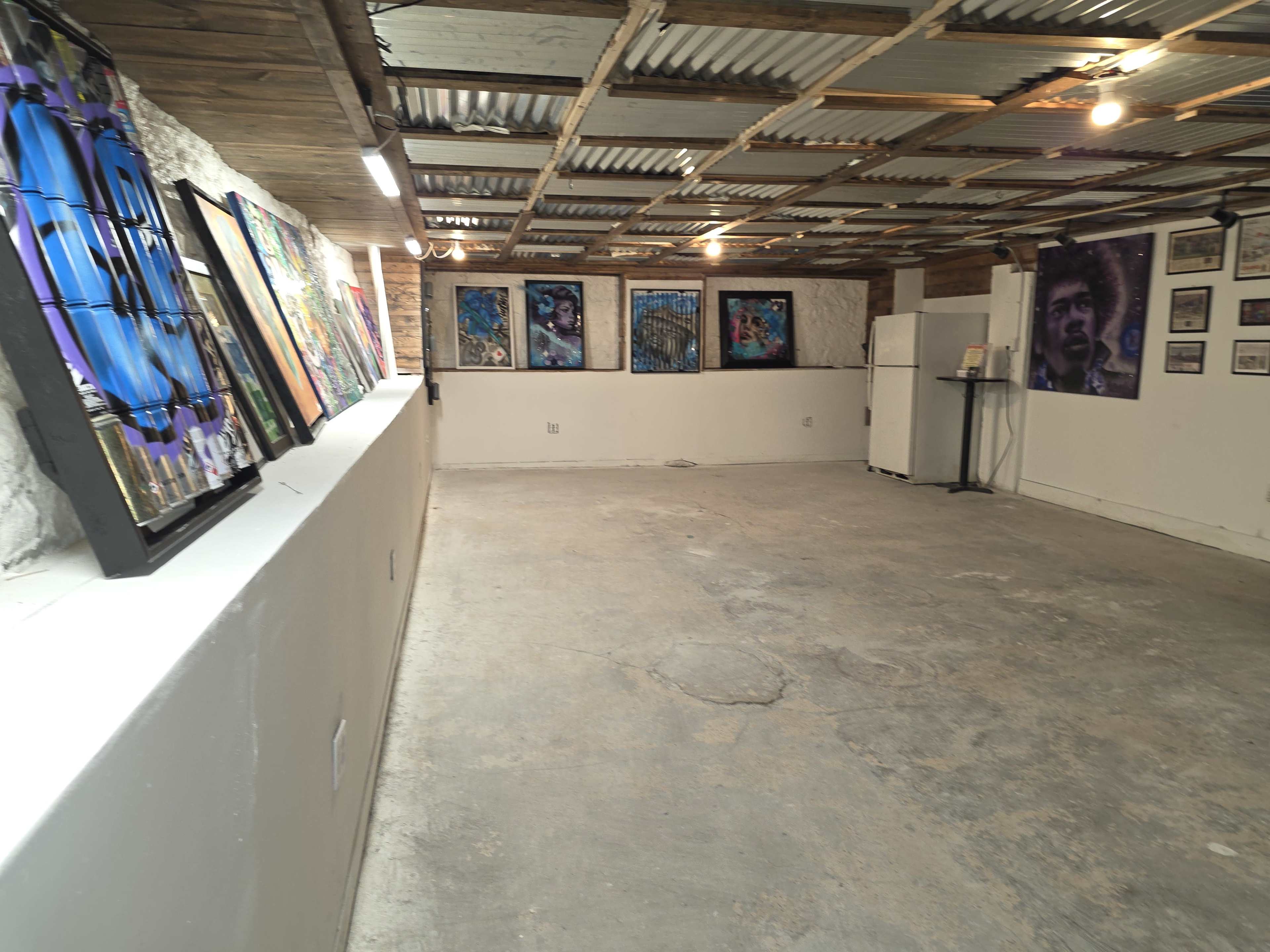 The image shows a spacious, empty room with concrete flooring, decorated with various framed artworks on the walls, and a refrigerator in one corner.