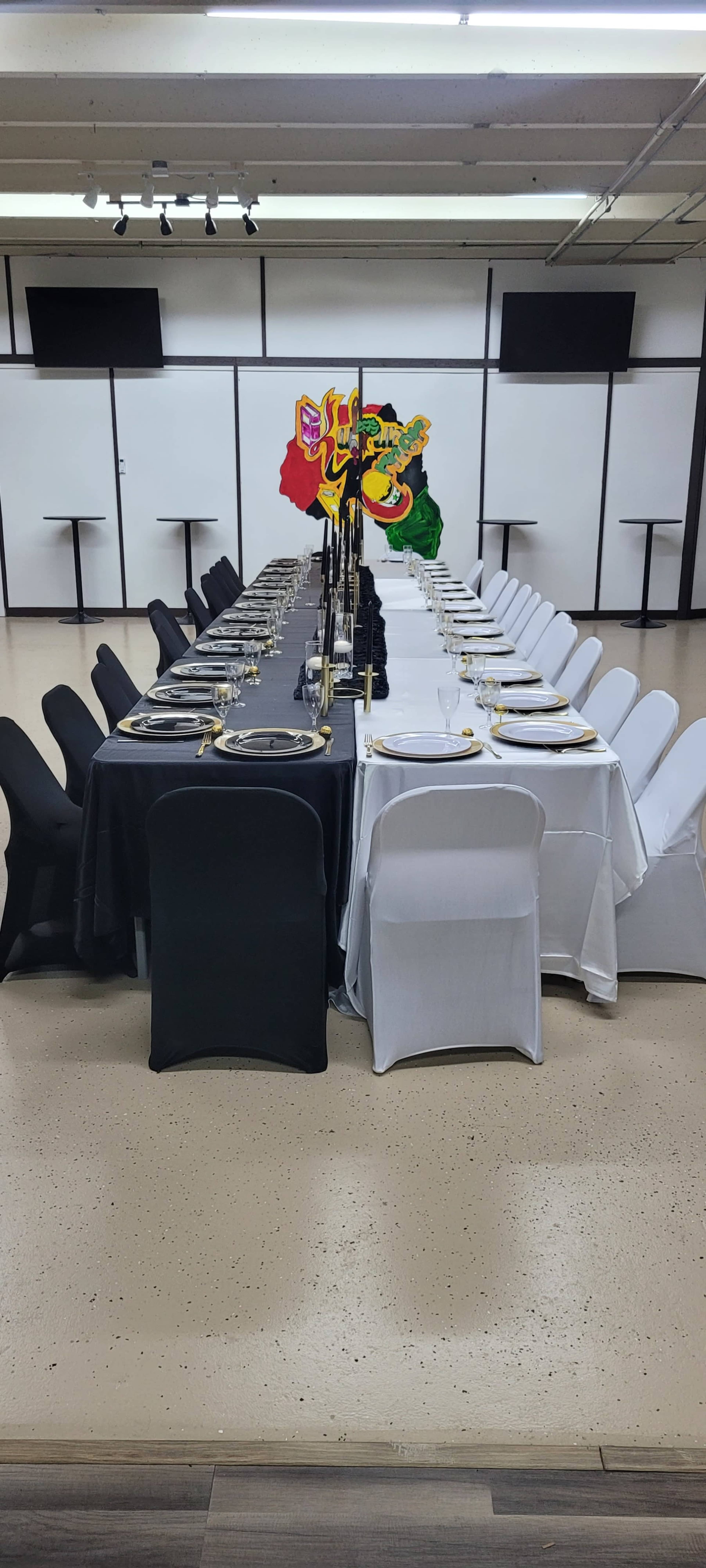 A long dining table is set with black and white tablecloths, plates, and glasses, featuring a decorative centerpiece, in a spacious room with minimalist decor.
