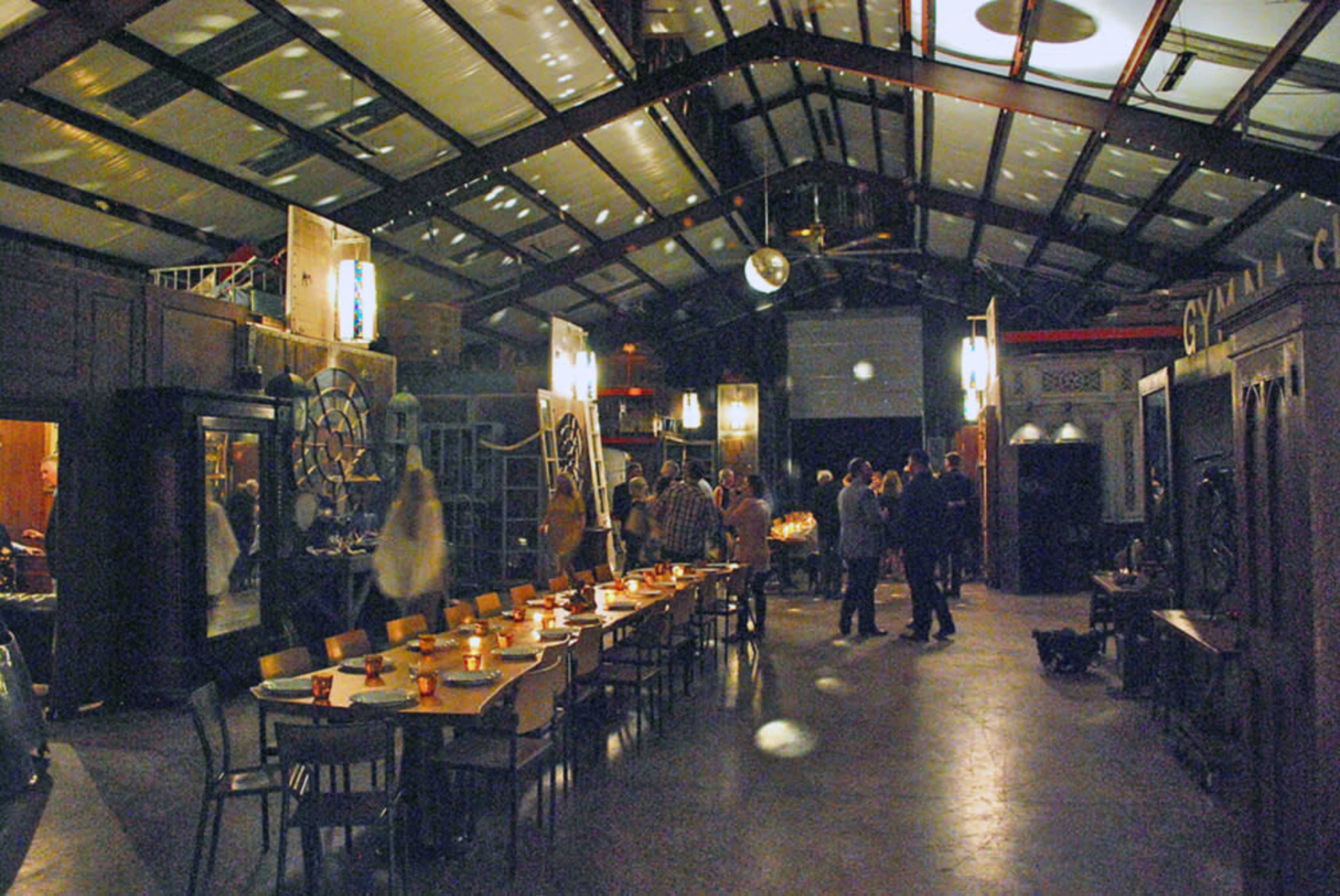 A spacious, dimly lit indoor venue features a long table set for dining, surrounded by guests mingling amid decorative lighting and wooden paneling.