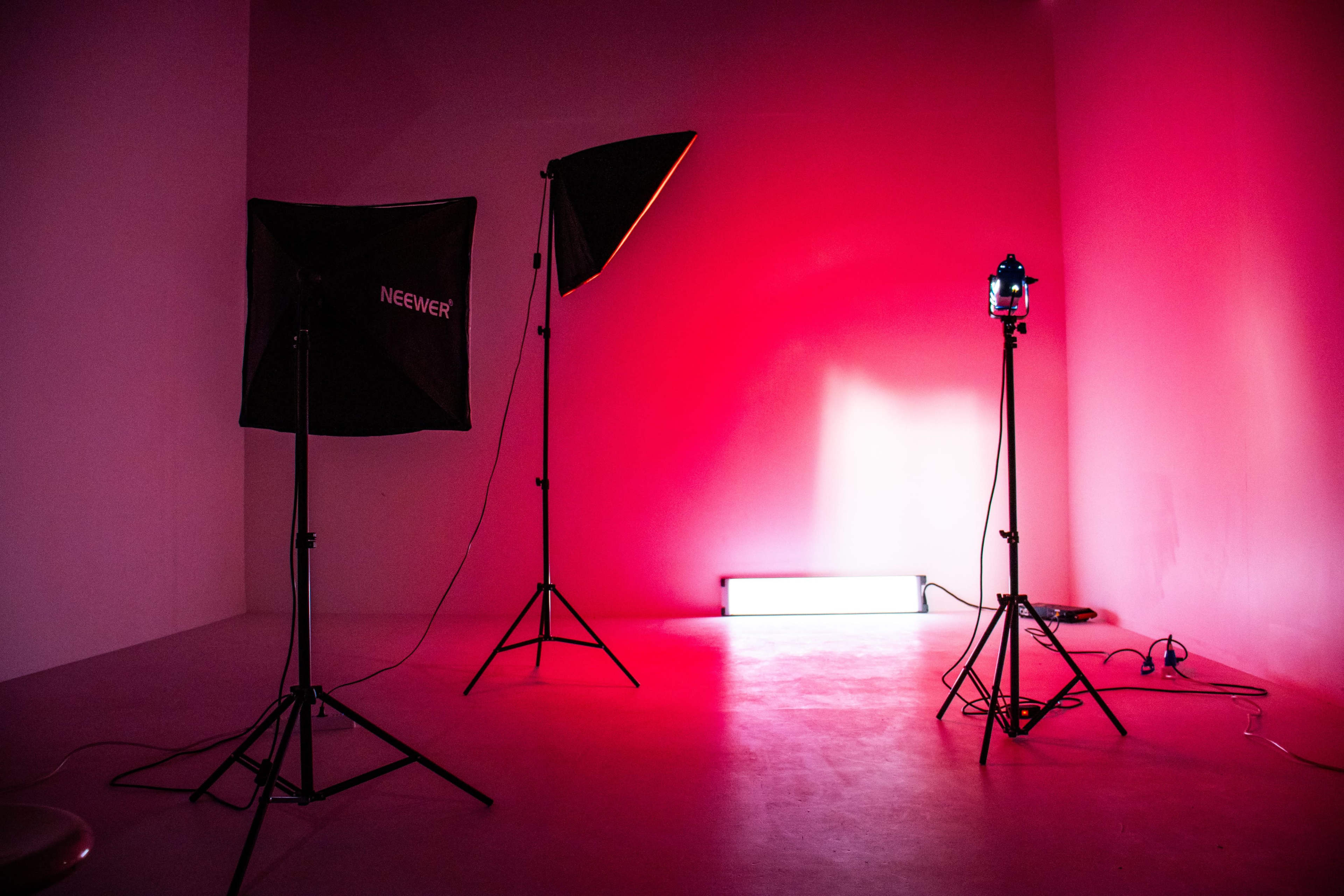 The image shows a sparse studio setup with two softbox lights and a neon light against a pink backdrop.