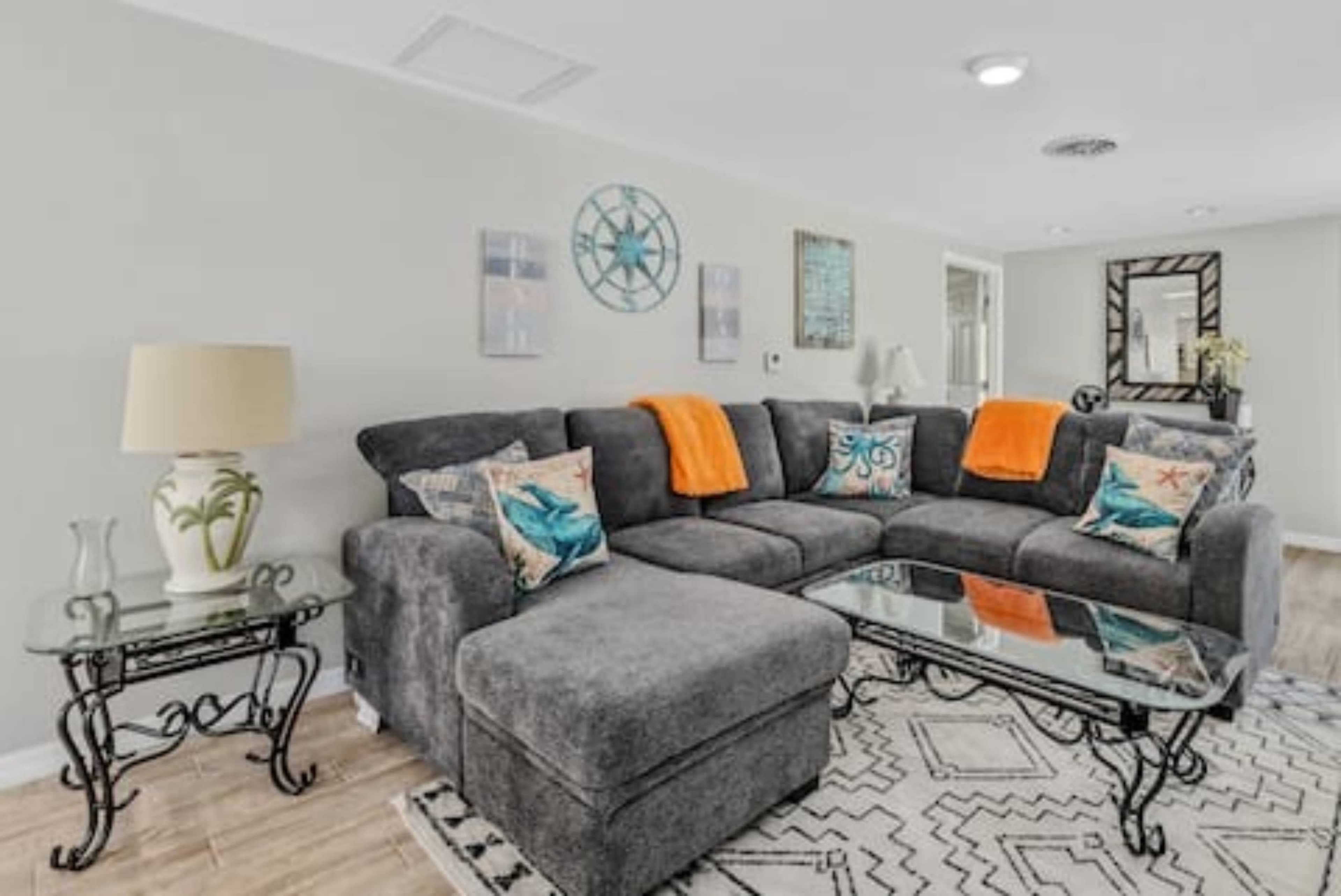 The image shows a modern living room with a gray sectional sofa adorned with orange throw blankets, a glass coffee table, and decorative pillows featuring aquatic themes.