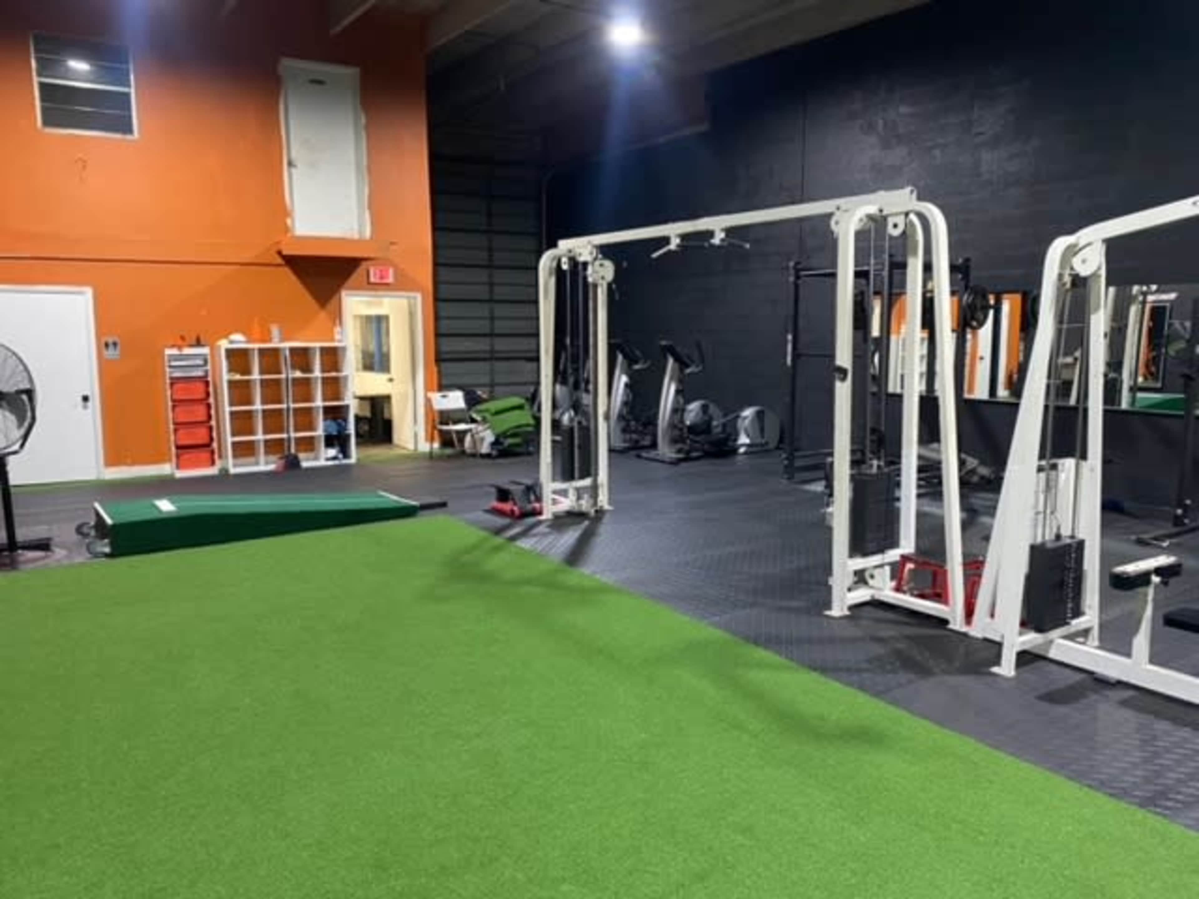 A gym interior featuring a turf area, workout equipment, and mirrors against a black wall with an orange accent wall.