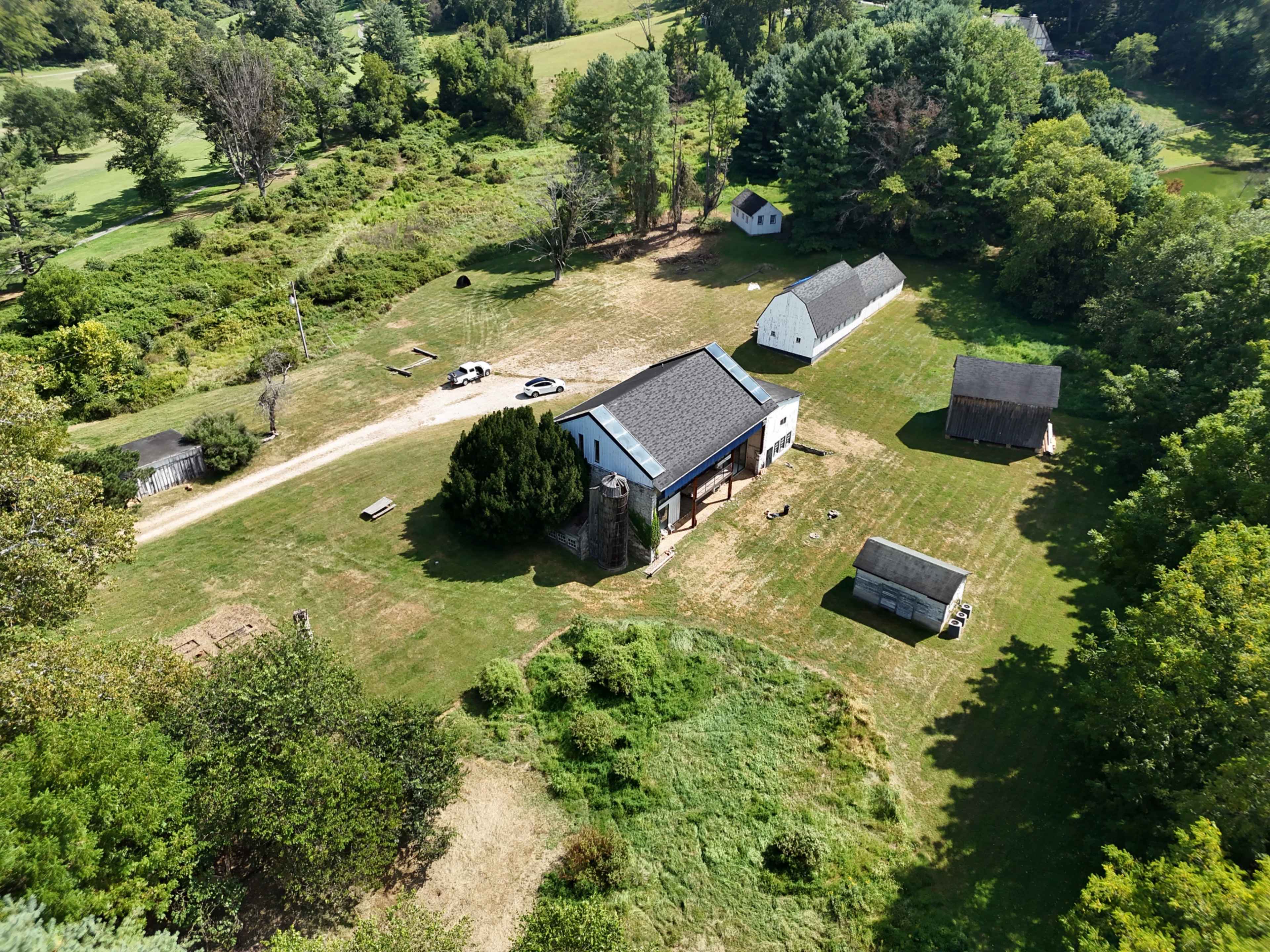 The image shows an aerial view of a rural property featuring several buildings, including a main house with a dark roof, a couple of smaller outbuildings, and open green spaces surrounded by trees.