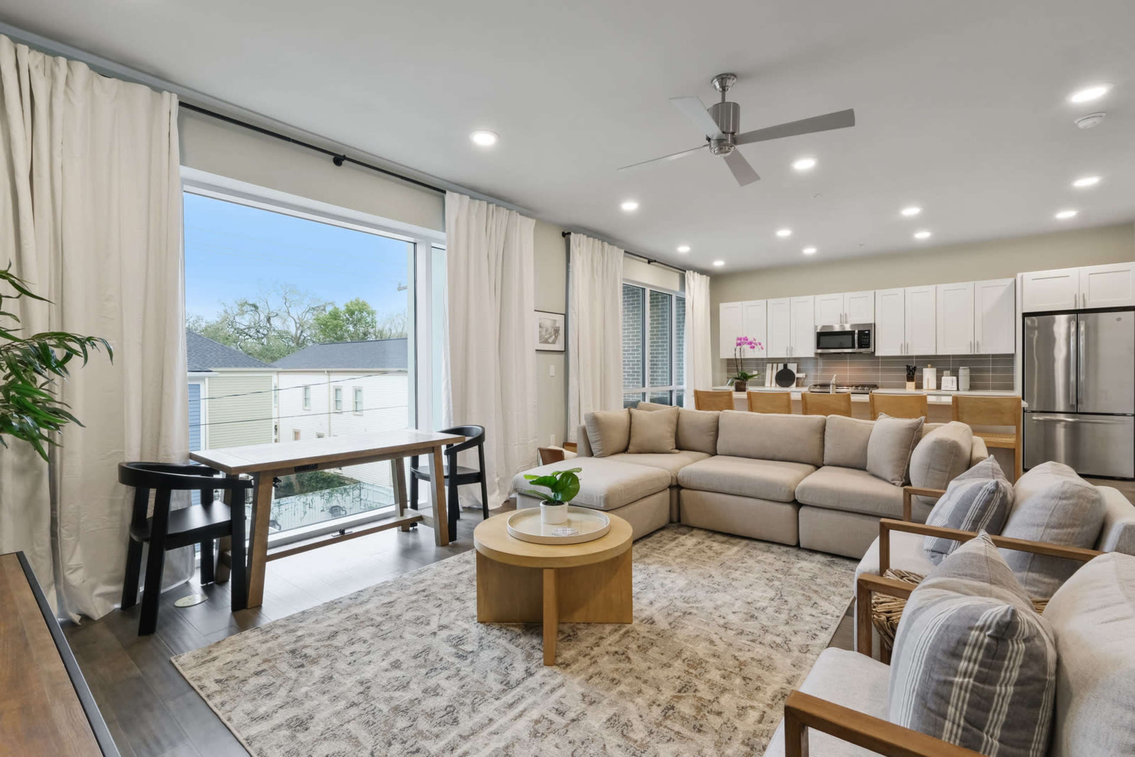 A modern living room features a large sectional sofa, a round coffee table, and a spacious kitchen in the background, with large windows allowing natural light to enter the space.