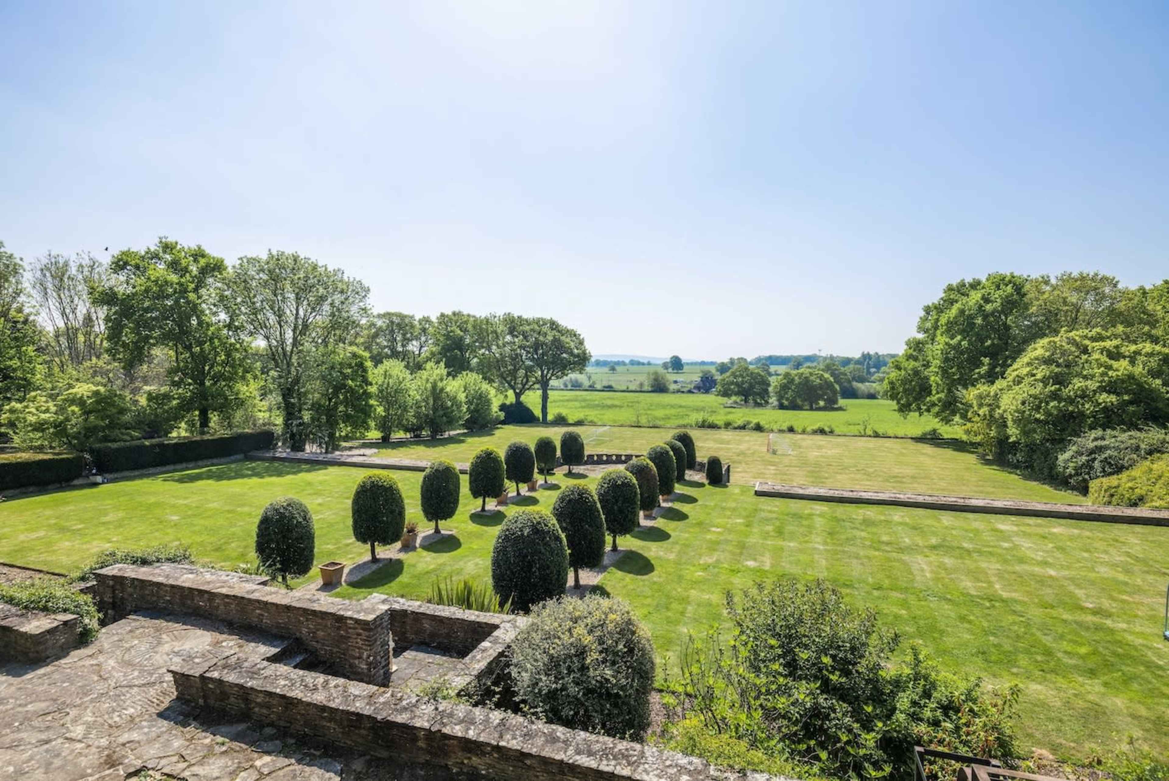 The image shows a well-maintained garden featuring neatly trimmed hedges arranged in rows, with a vast green field and trees visible in the background under a clear blue sky.