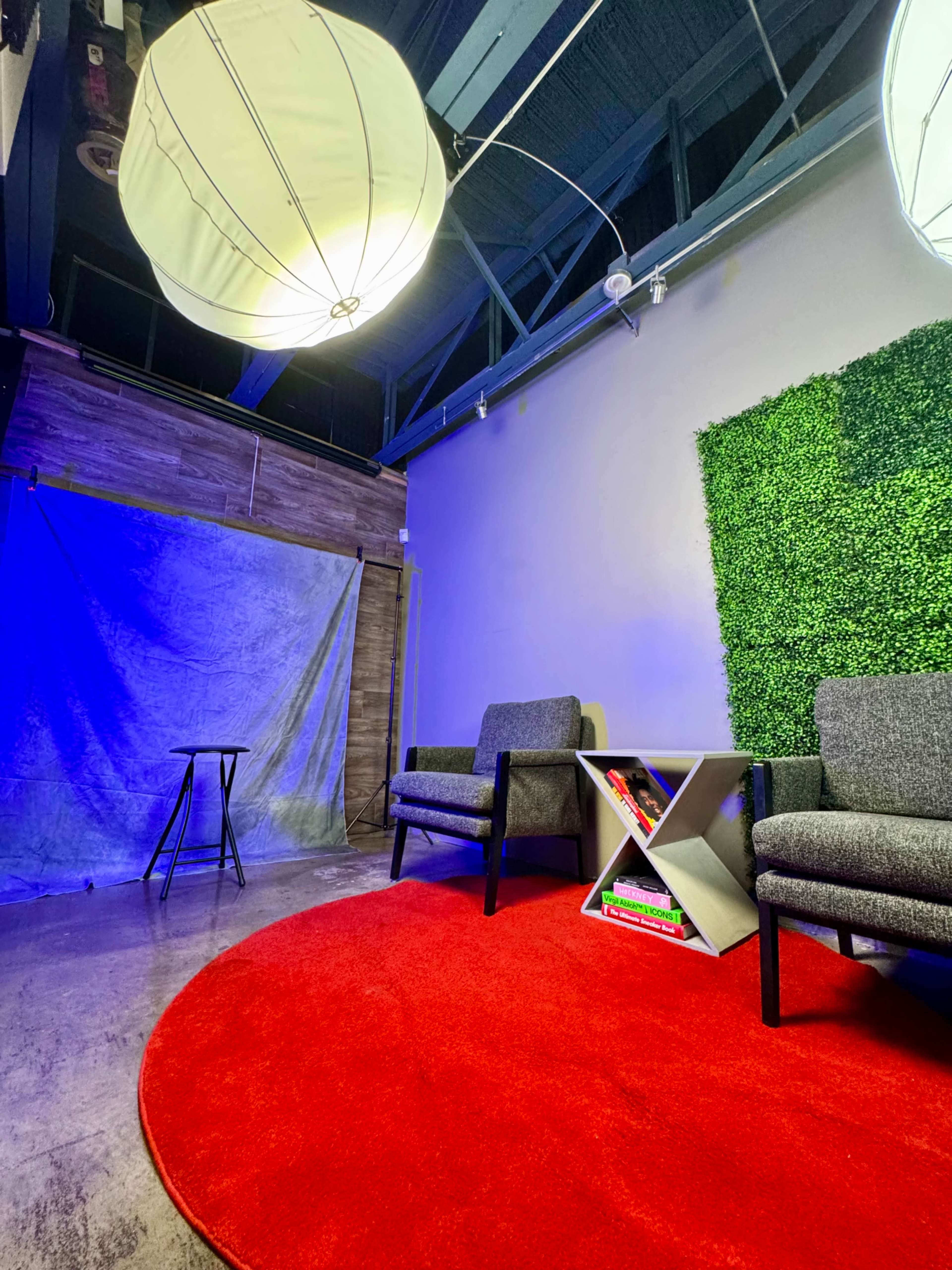 The scene features a modern interior with two gray chairs on a red circular rug, a small table beside them, a green wall of faux foliage, and a backdrop near a blue-lit area.