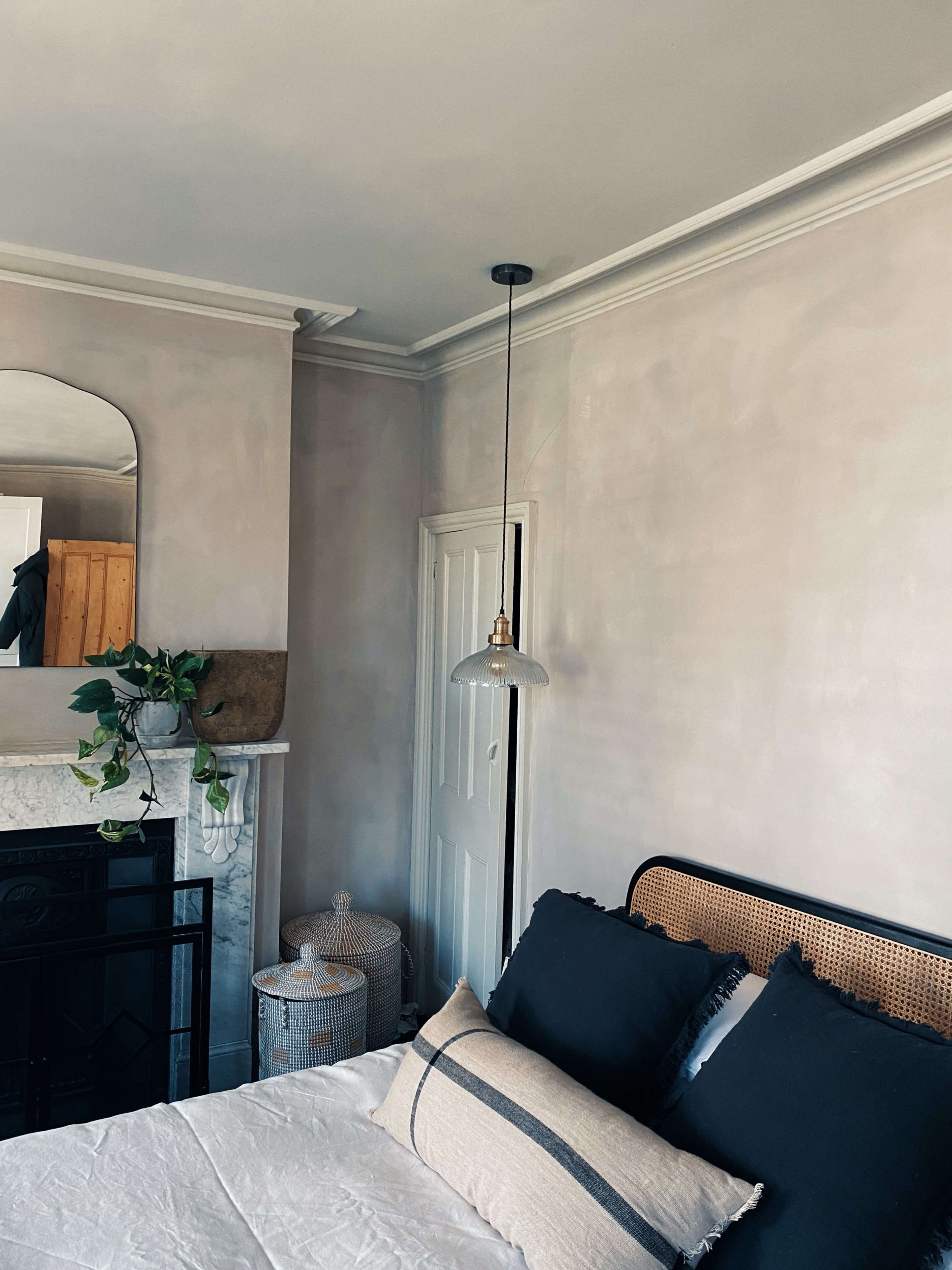 A bedroom featuring a bed with dark pillows, a pendant light hanging above, a decorative mirror on the wall, and a visible doorway and fireplace.