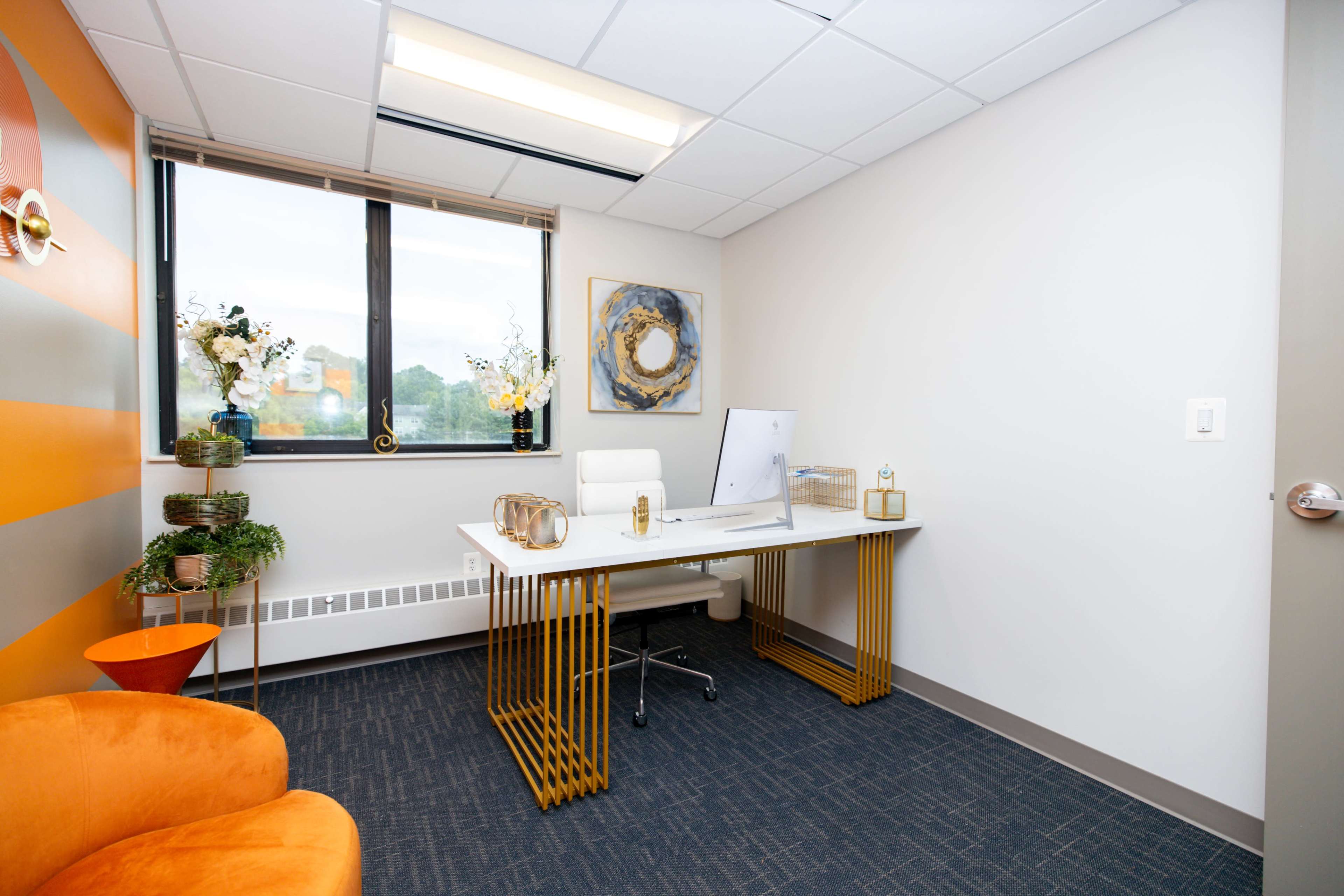 The image shows a modern office space featuring a white desk with a computer, an orange chair, and decorative plants on the windowsill.