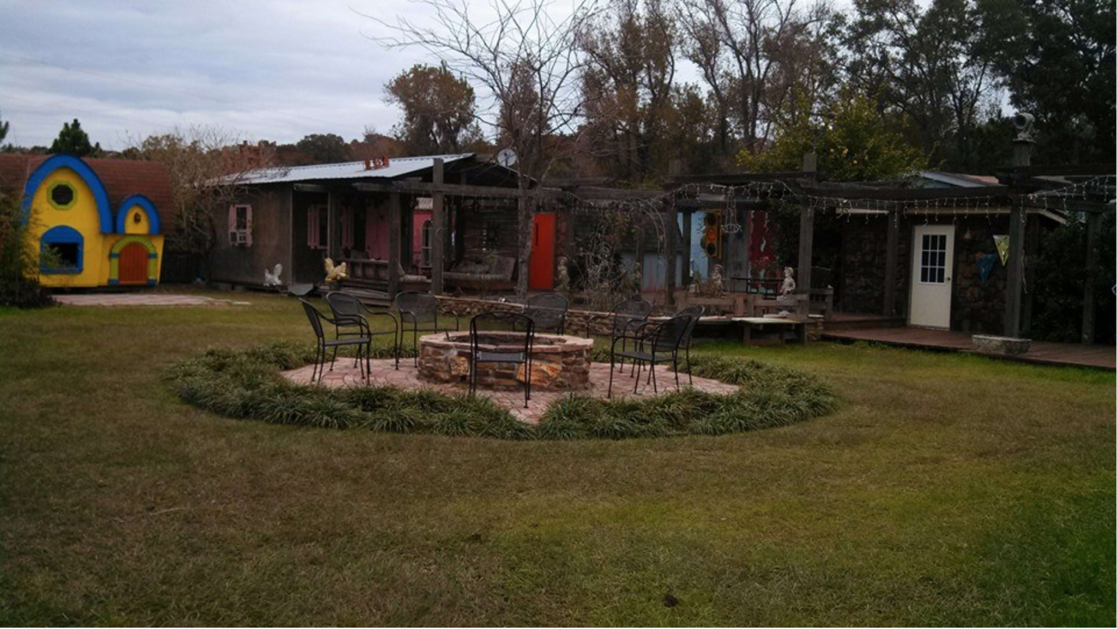 A grassy outdoor space with a stone fire pit surrounded by chairs, a colorful playhouse, and several rustic buildings in the background.