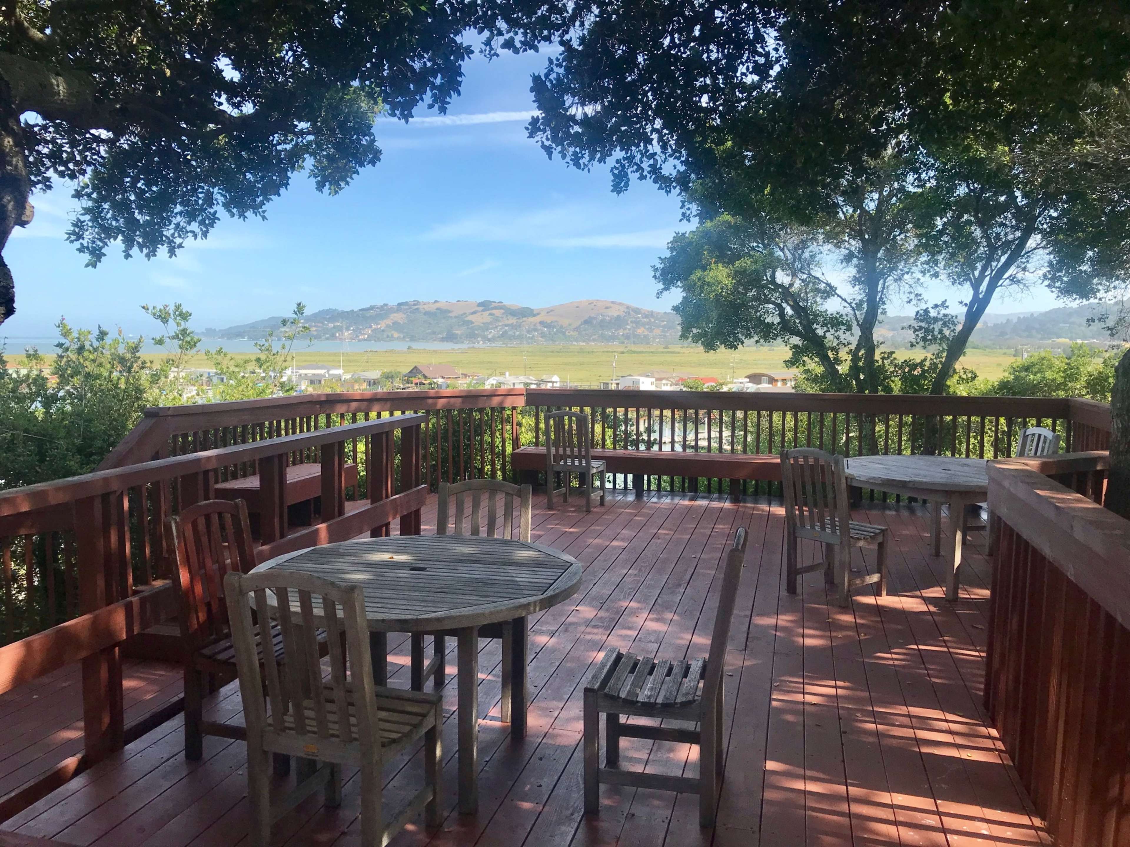 A wooden deck with several tables and chairs overlooks a landscape of hills and trees under a clear sky.