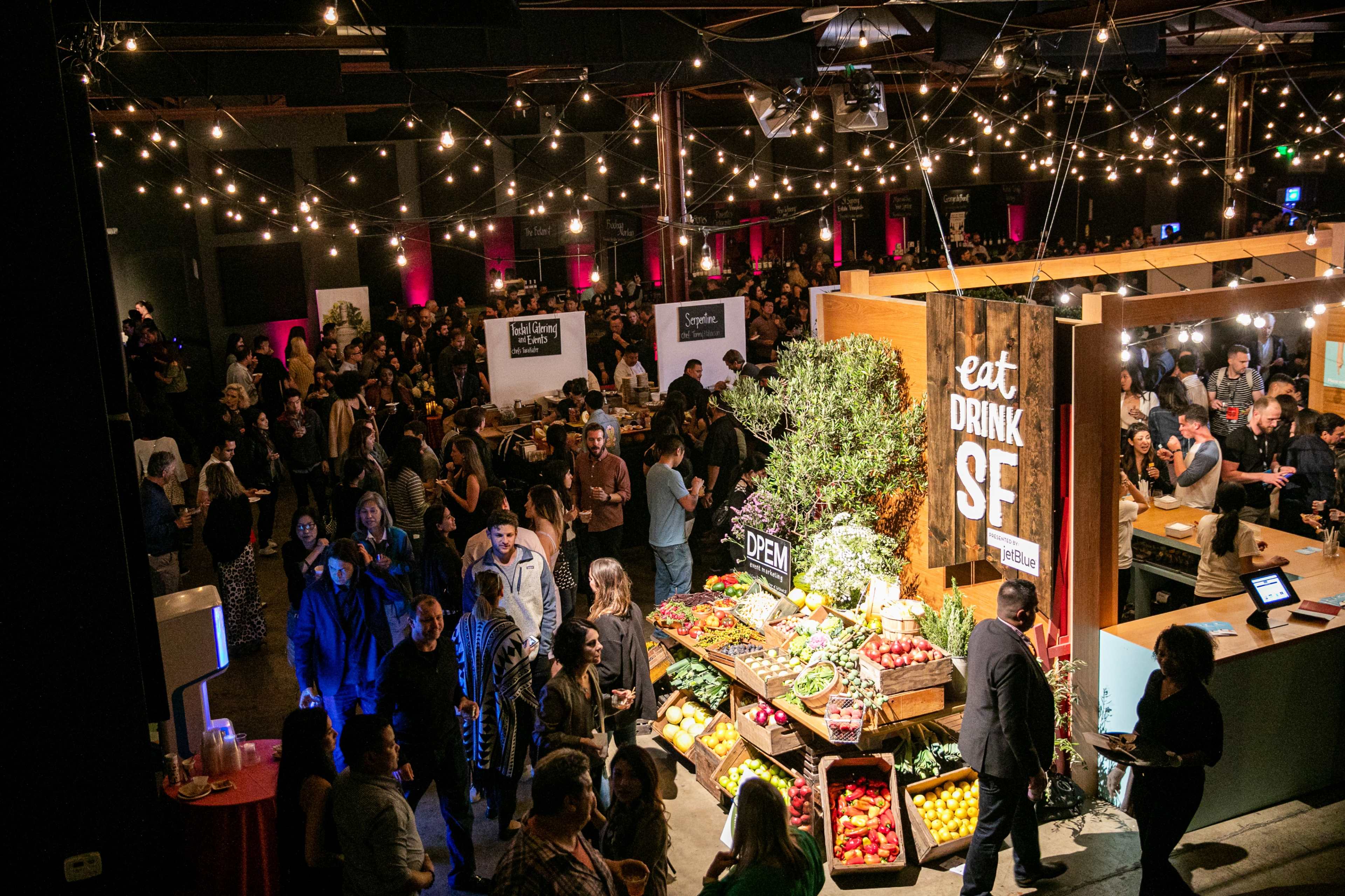 A crowded event space features booths showcasing food and drinks, with stalls decorated with fresh produce and string lights overhead.