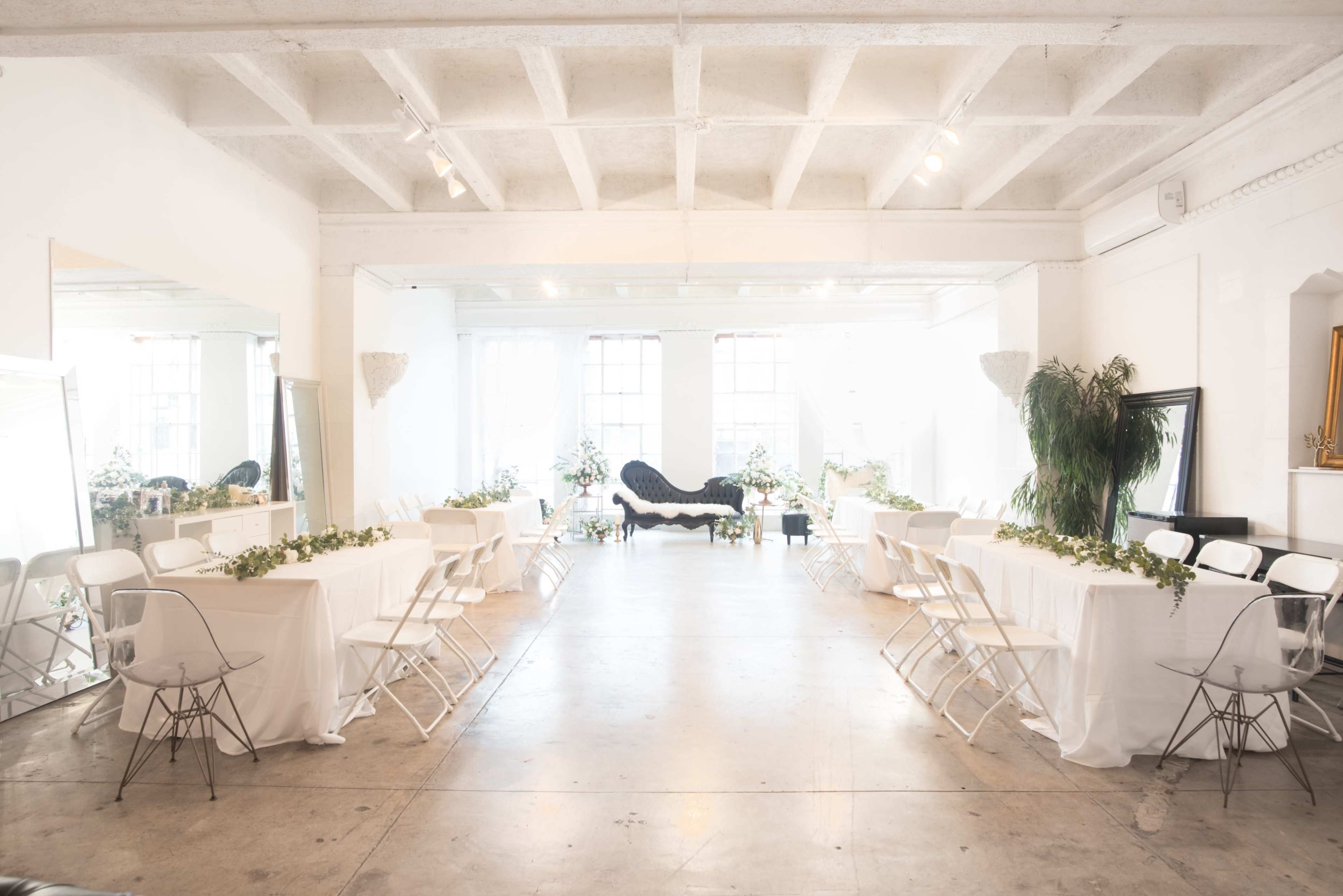 A bright, modern event space features long tables covered in white linens, surrounded by chairs, with greenery and decorative mirrors enhancing the atmosphere.