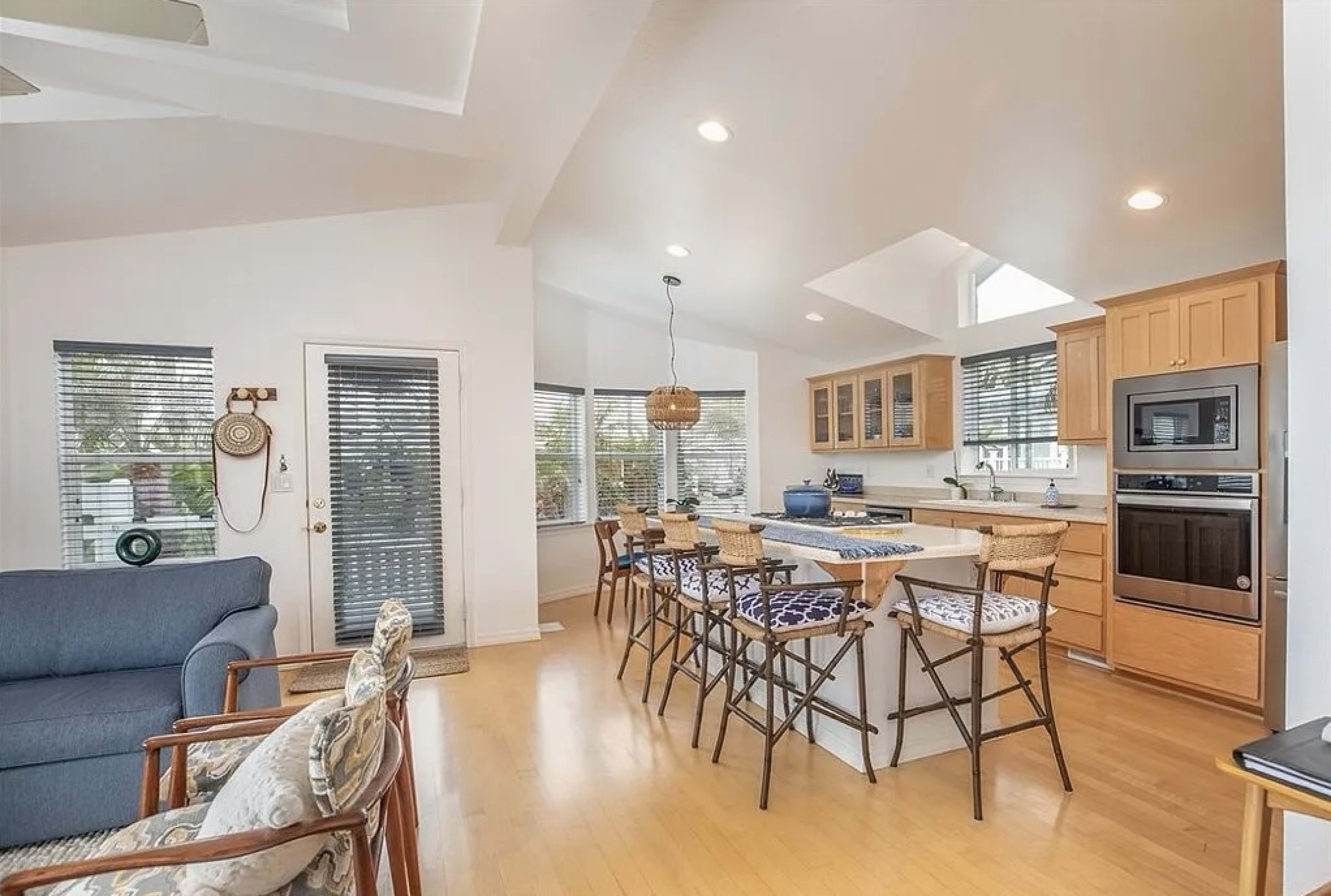 A well-lit kitchen and living area feature a central island with seating, wooden cabinetry, and large windows.