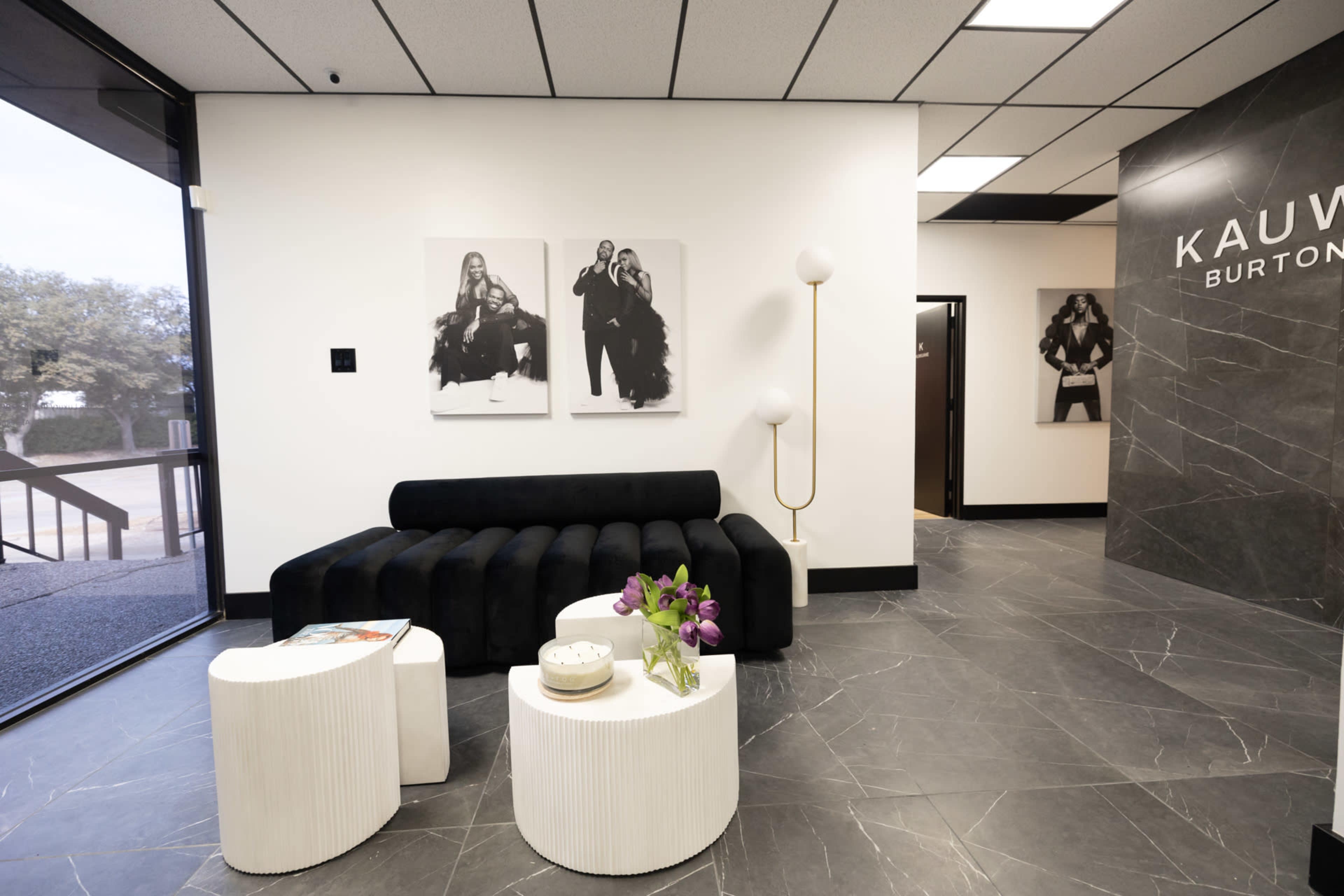 The image shows a modern office reception area with a black couch, round white tables, and framed black-and-white photographs on the walls.