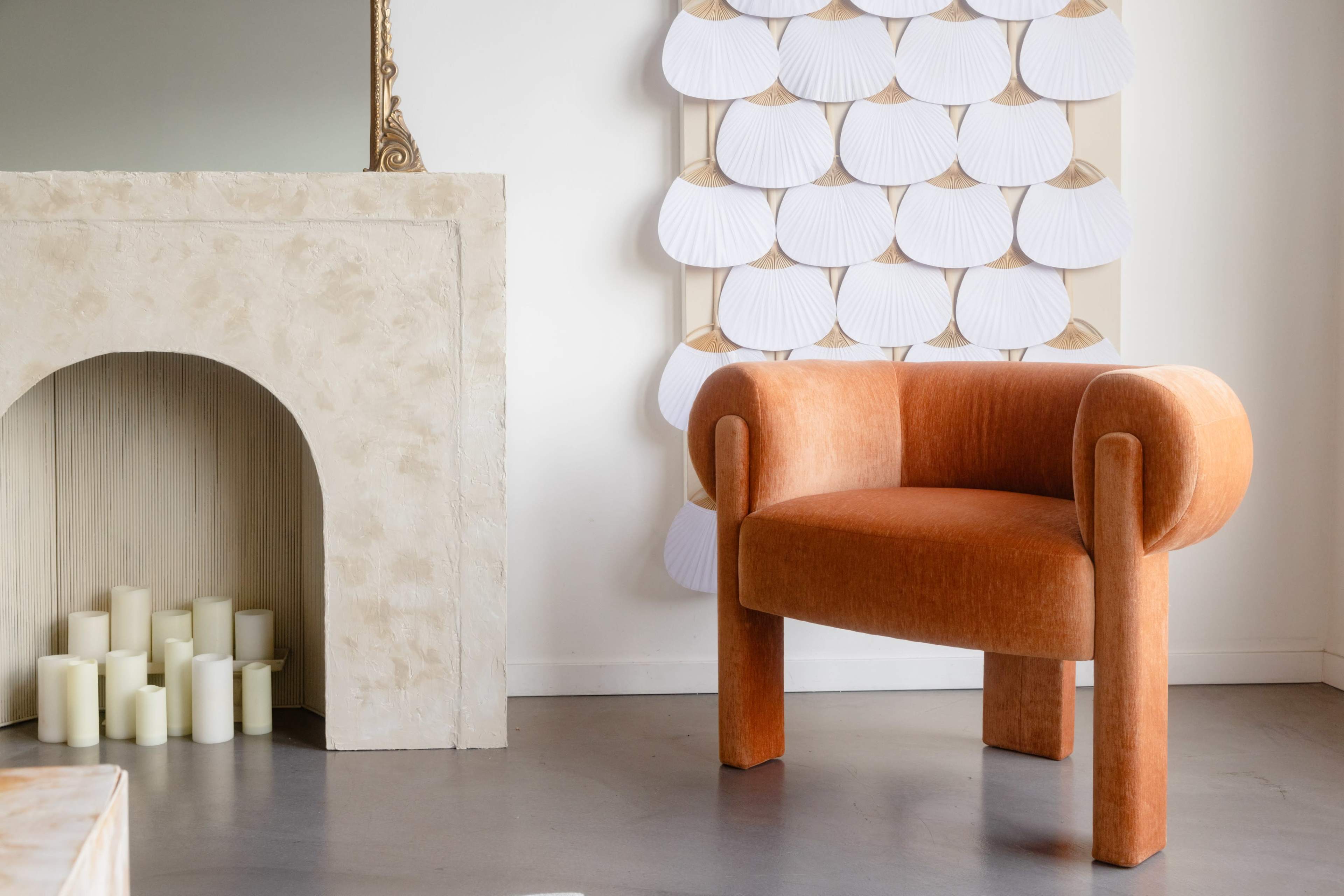 The image shows an orange upholstered chair next to a modern fireplace and a decorative wall art piece featuring white, petal-like shapes.