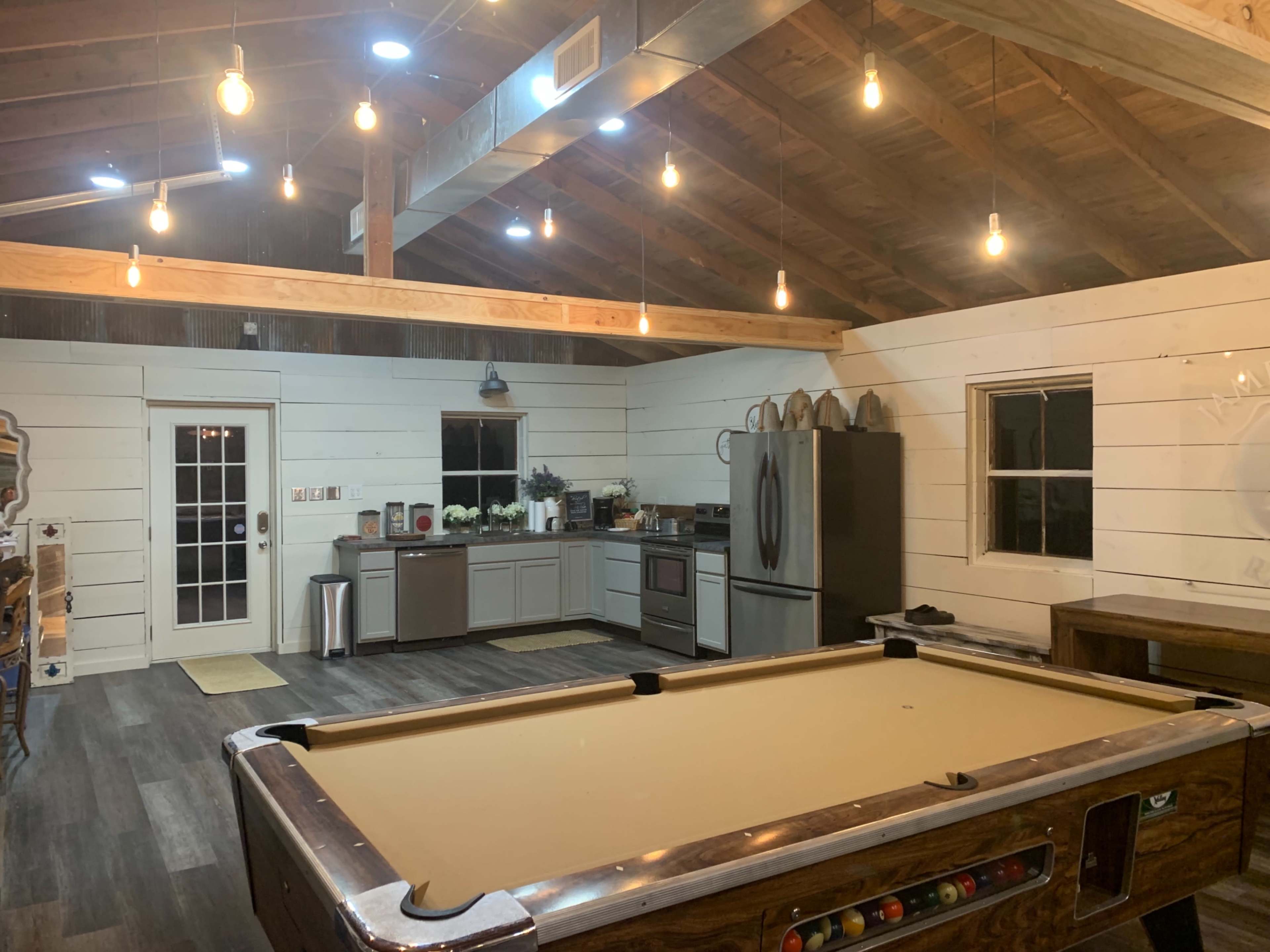 The image shows a spacious room with a pool table in the foreground, wooden beams on the ceiling, and a kitchen area with modern appliances in the background.
