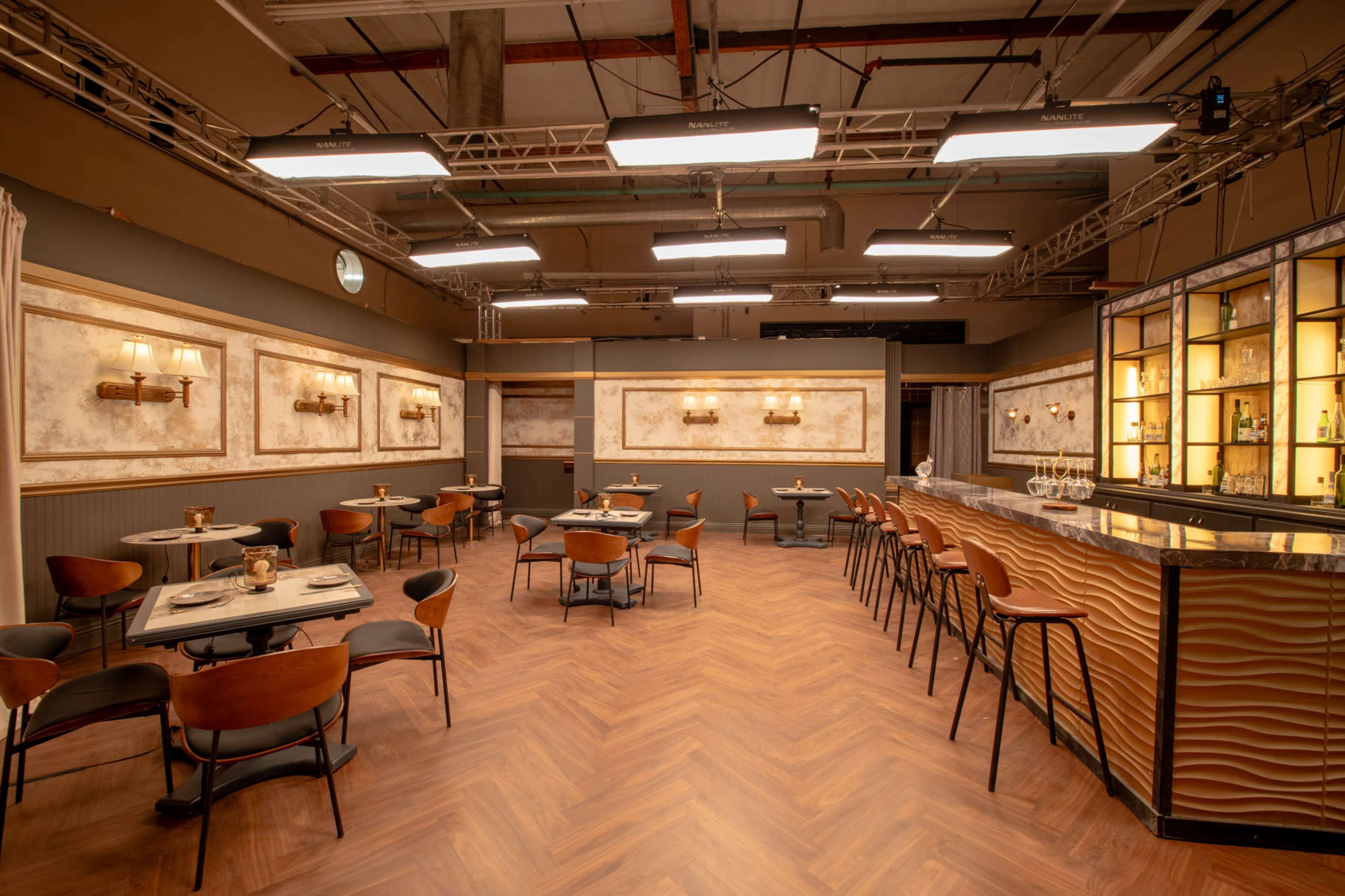 The image shows a modern restaurant interior with wooden floors, round tables, and a long bar featuring shelves of glassware.