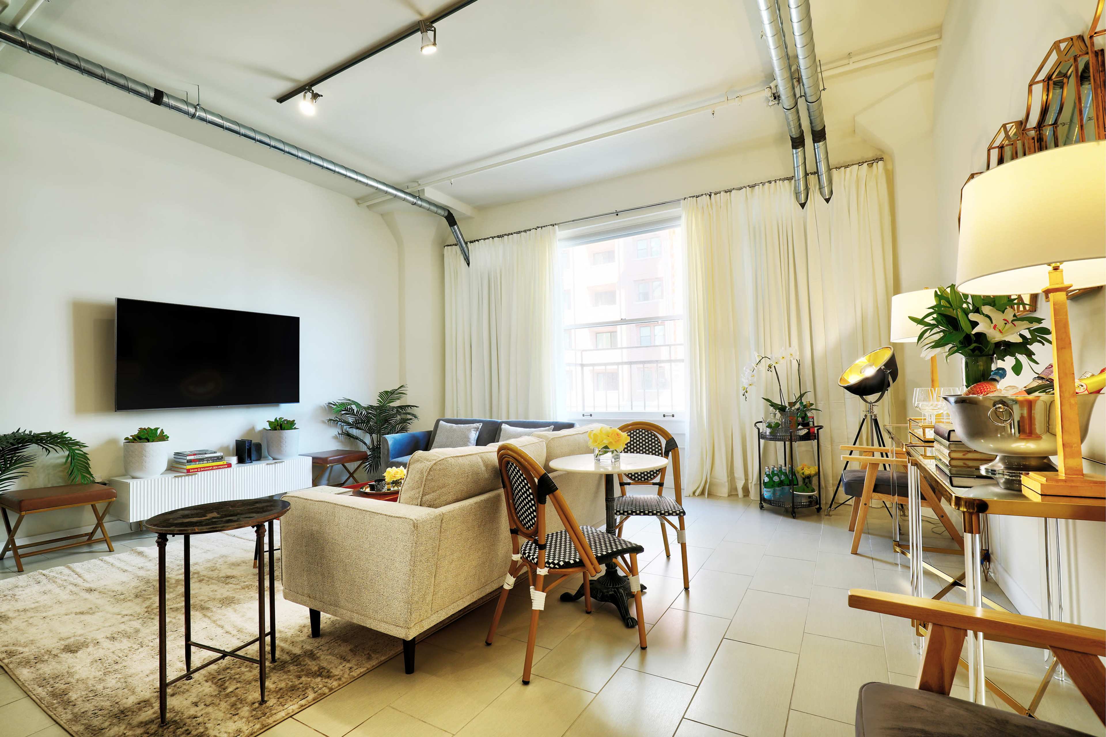 The image shows a modern living room with a large television mounted on the wall, a comfortable seating area, and decorative plants.