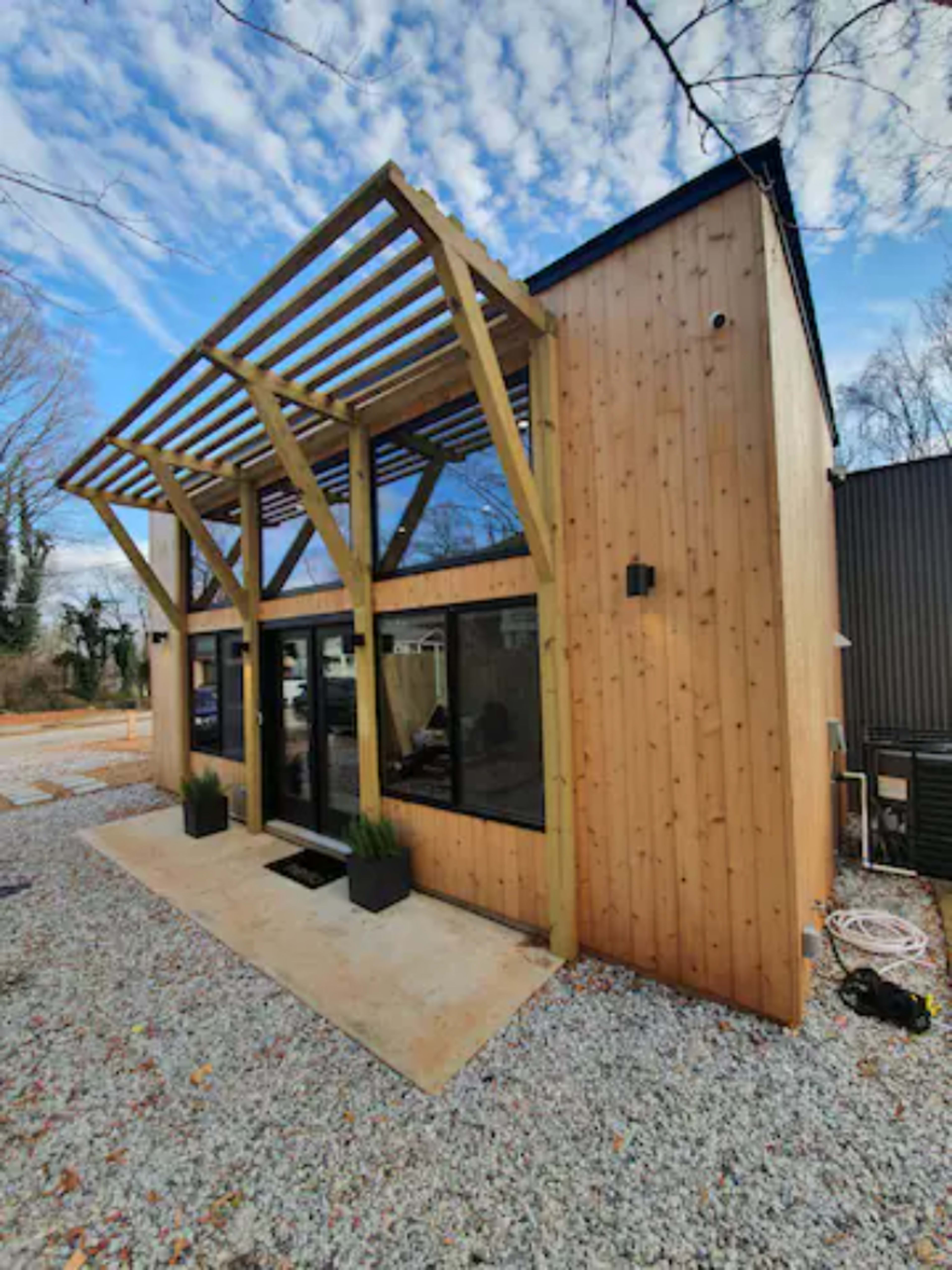 A modern wooden house with a sloped roof and large windows, set on a gravel surface.