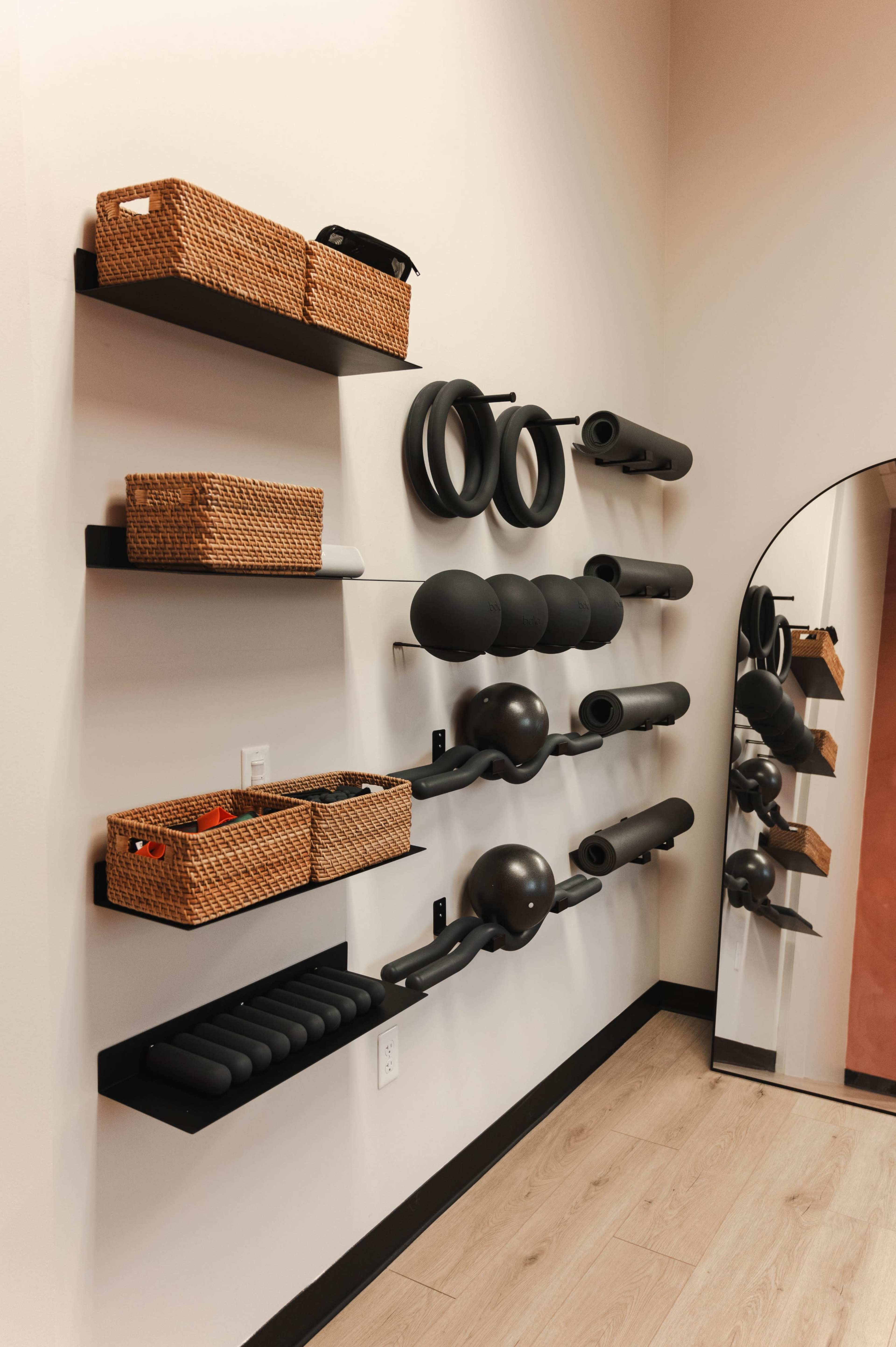 The image shows a wall with black shelves displaying exercise equipment and woven baskets for storage, alongside a large mirror.