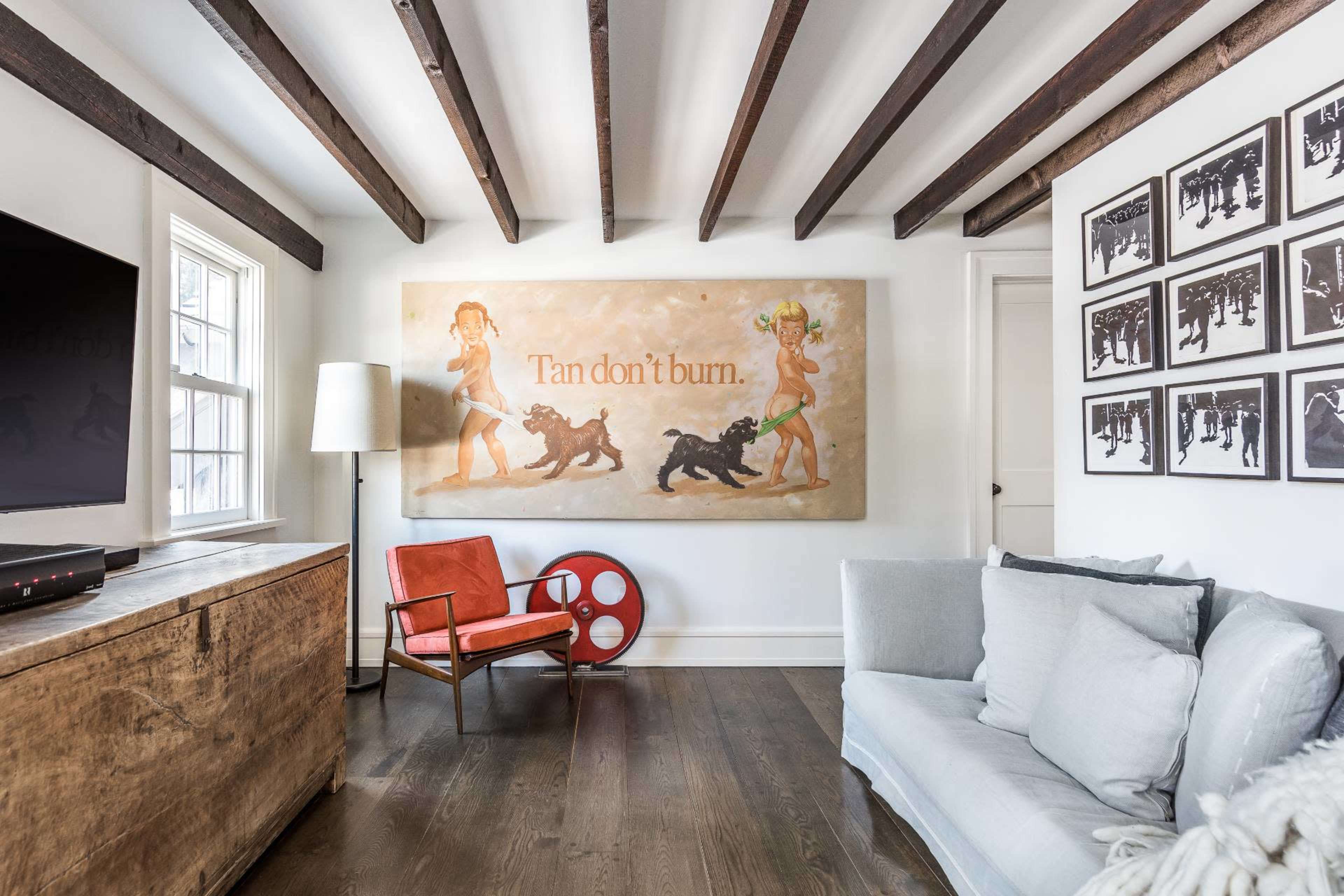 The room features a light-colored sofa, a red chair, a wooden cabinet, and a large wall art piece depicting two figures and dogs, all beneath exposed wooden beams.