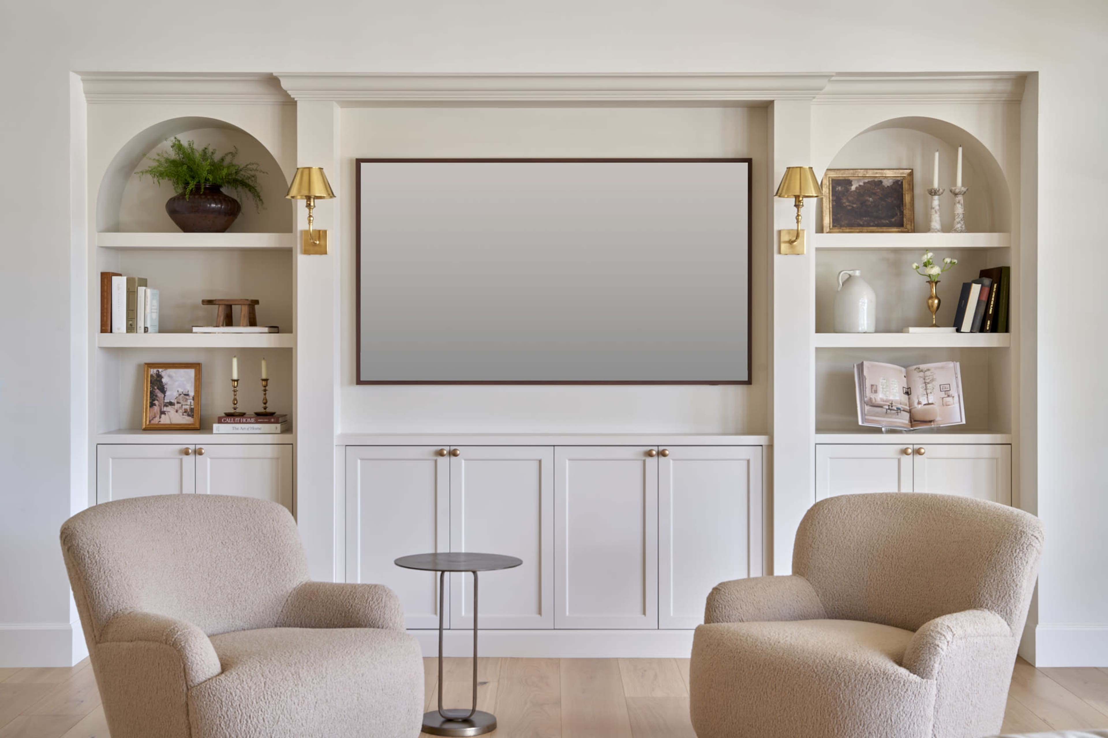 The image features a modern living room with two upholstered chairs facing a built-in shelf unit that includes framed photos, books, and decorative items, along with a flat-screen television mounted above a cabinet.