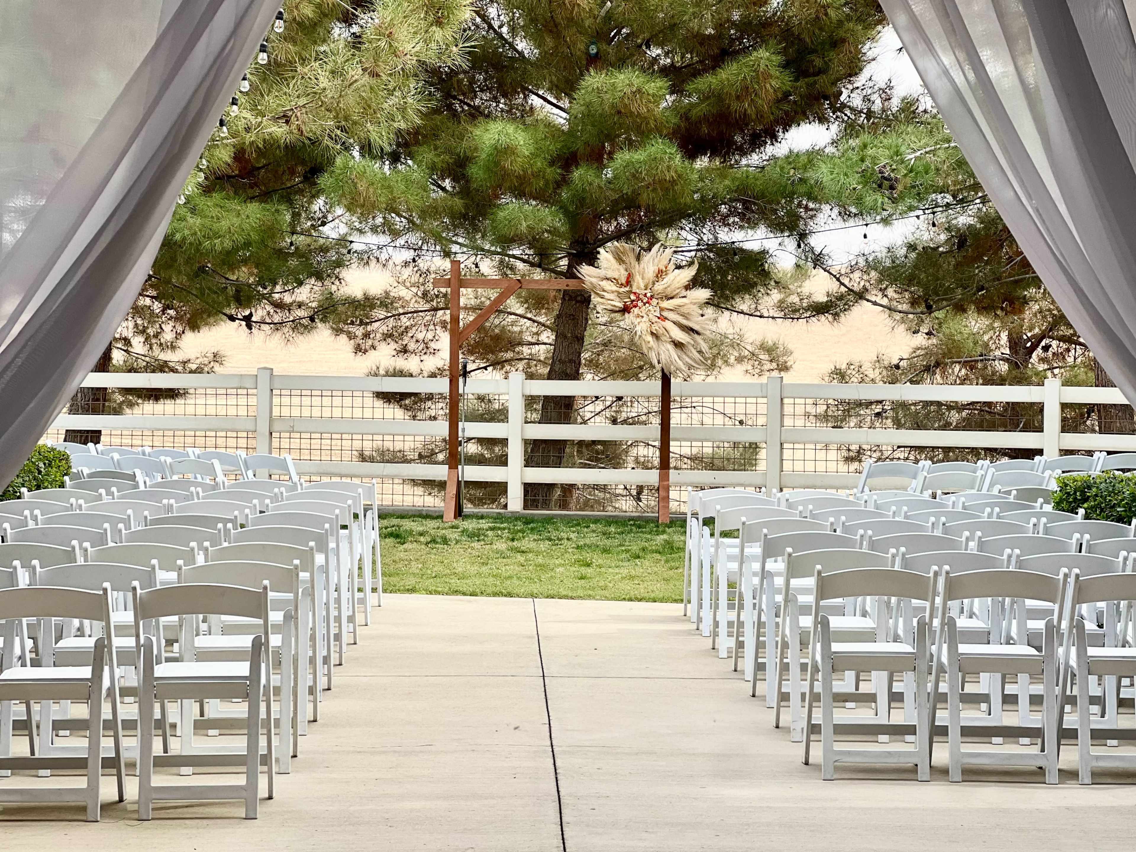 A wedding ceremony space is set up with rows of white chairs facing a decorative backdrop among greenery.