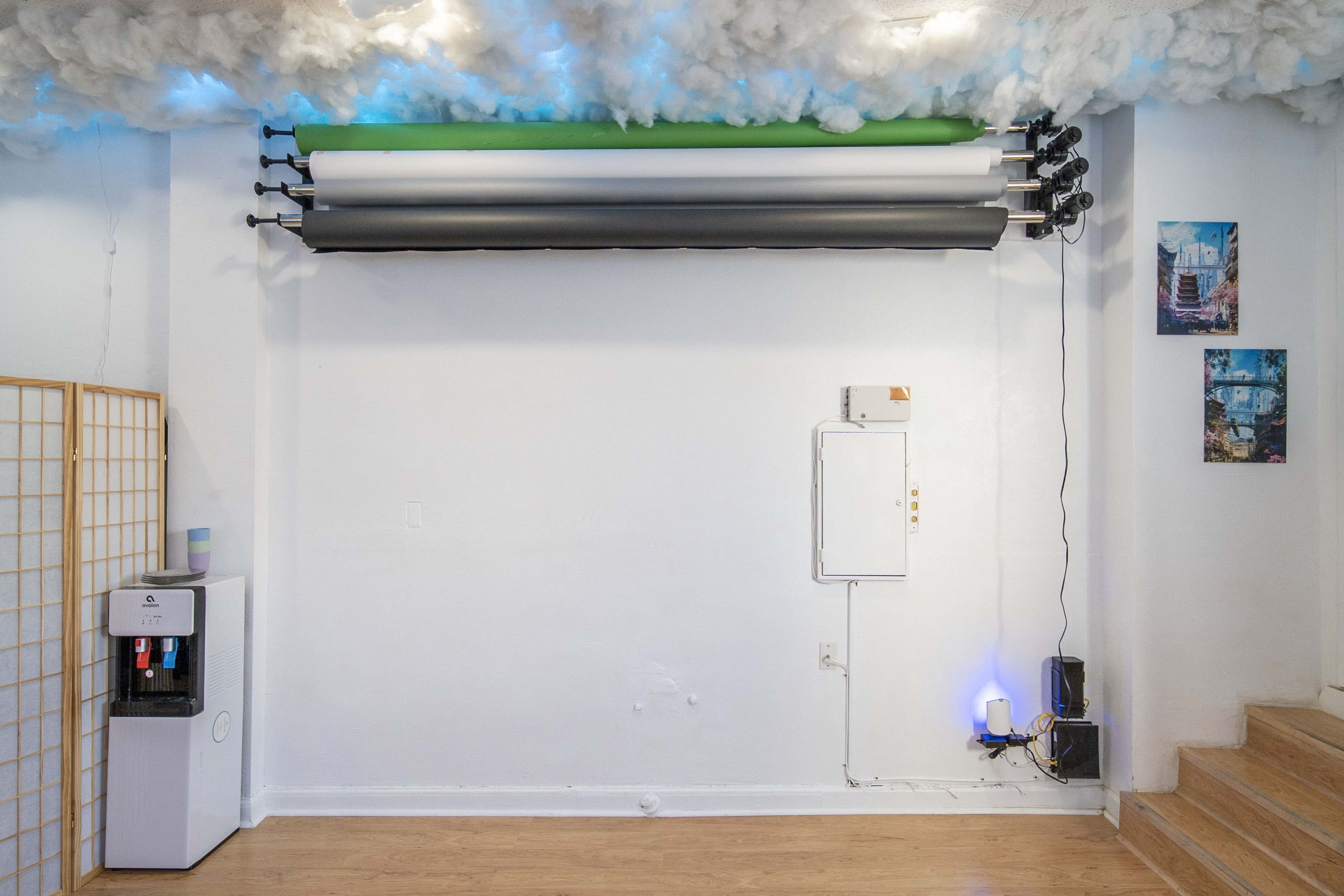 A studio space with a white wall, a water dispenser, a control panel, a set of rolled backdrops mounted on a rod, and two framed photos on the adjacent wall.