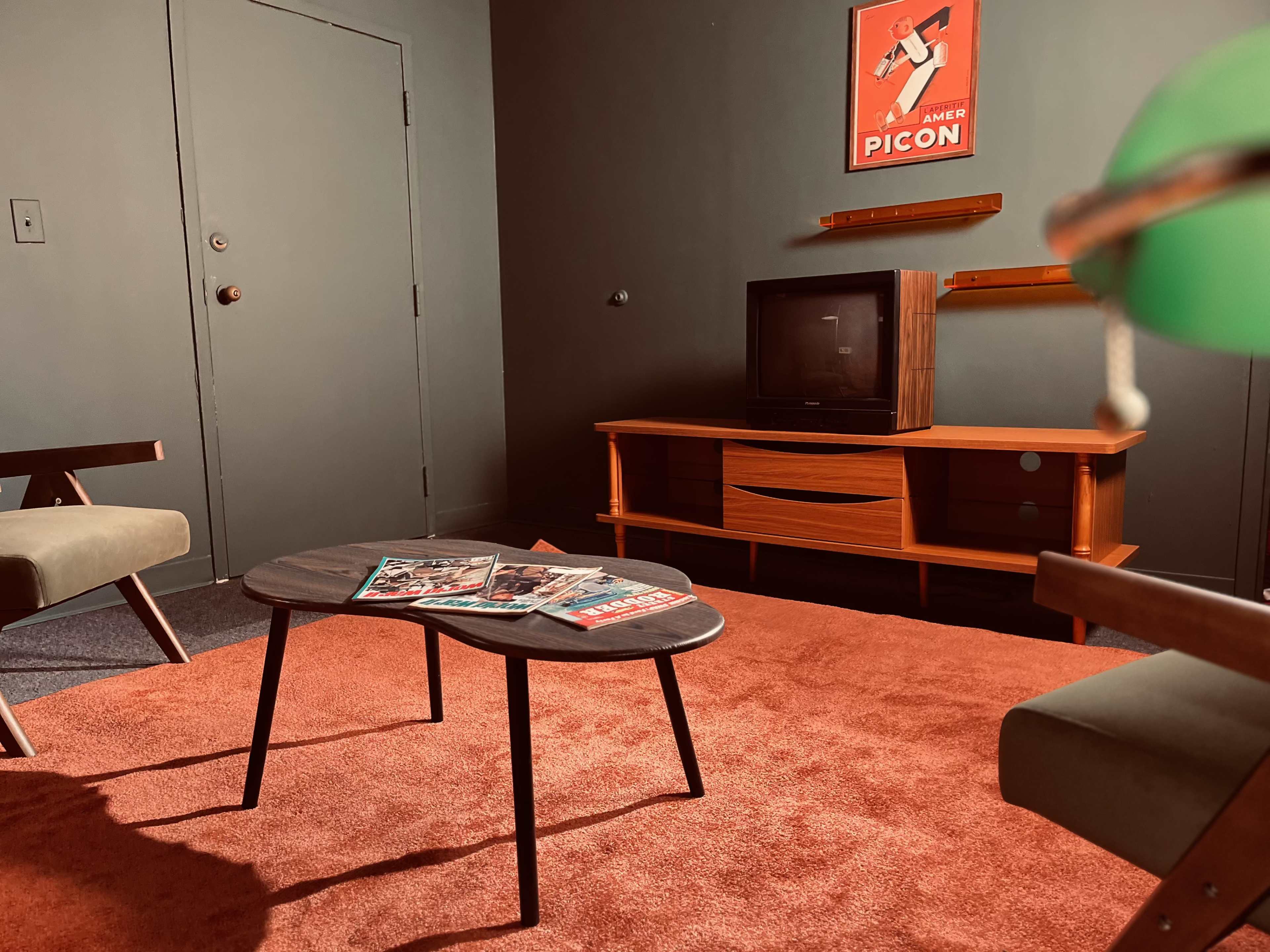 The image shows a retro-styled living room with a low table, two armchairs, an old television set, and a vibrant orange carpet.