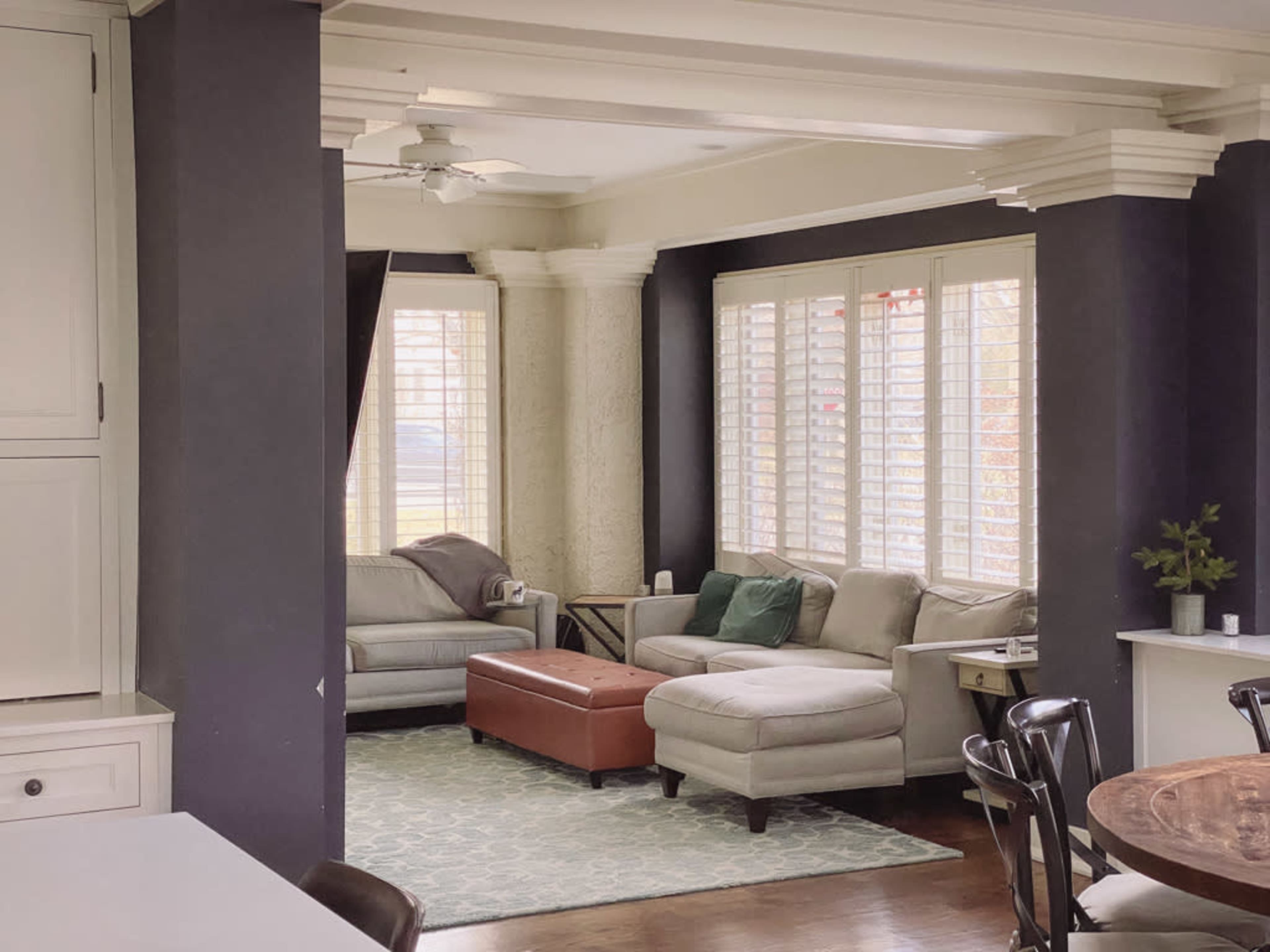 The image shows a living room with light-colored sofas, a brown ottoman, and large windows covered with white shutters.