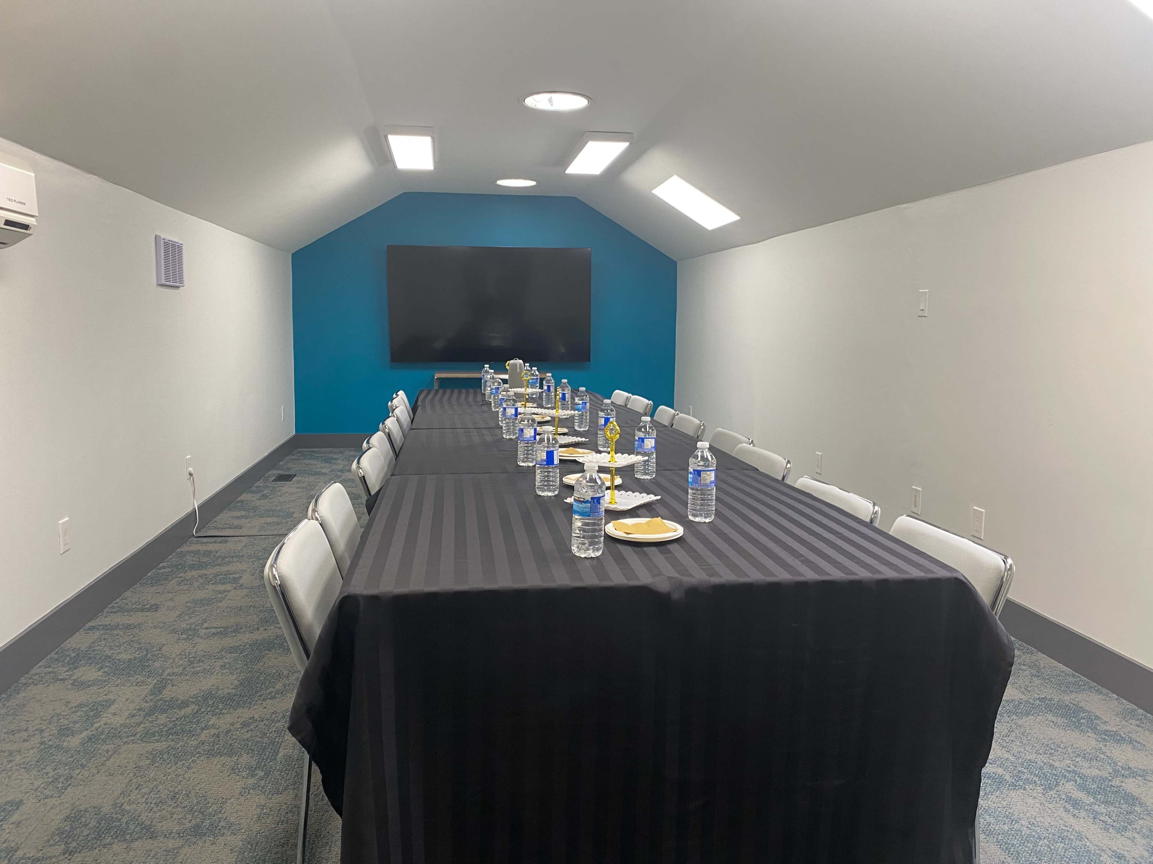 A conference room with a long table set with bottled water and plates, and a wall-mounted screen at the far end.