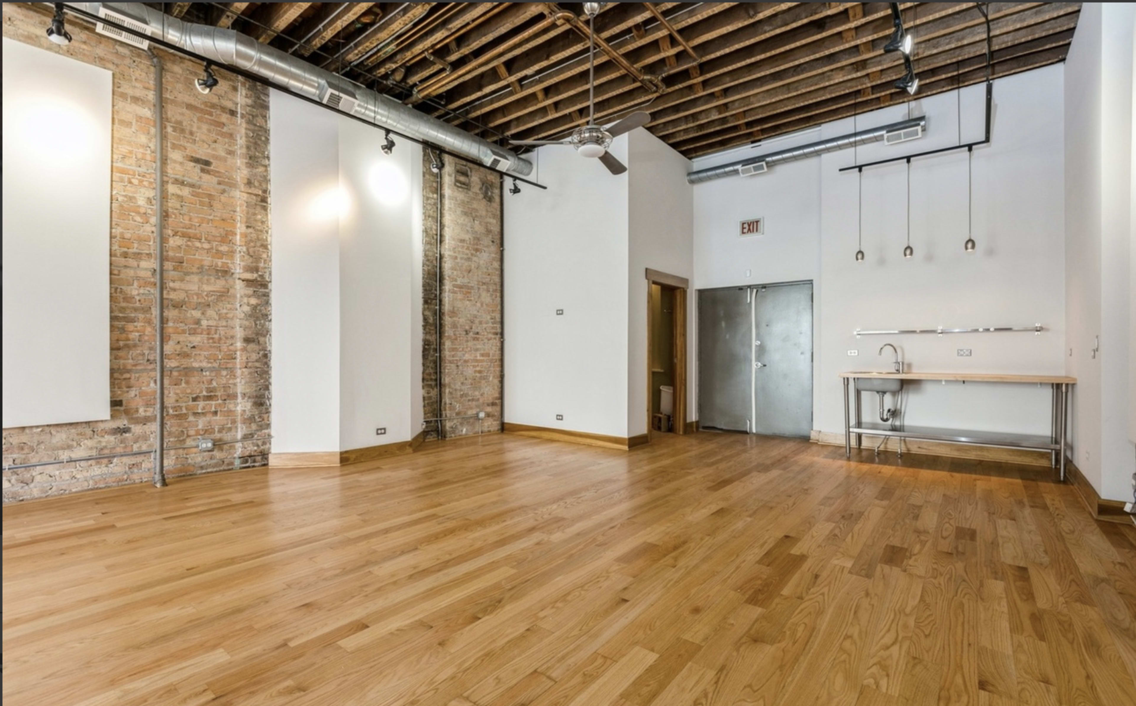 The image shows a spacious, minimalist interior with hardwood flooring, exposed brick walls, and a small kitchenette area.