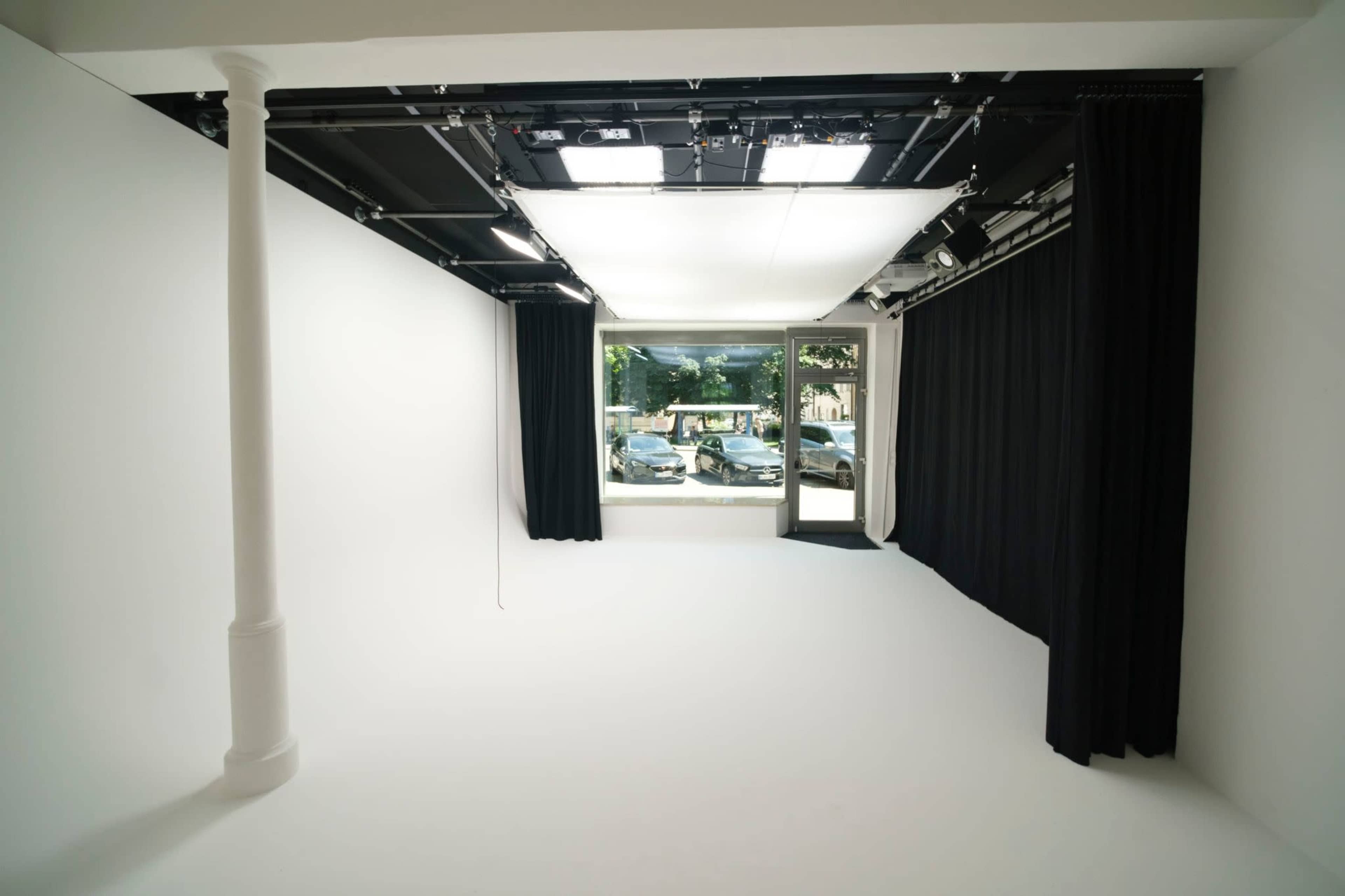 The image shows a spacious, minimalistic interior with white walls, a large window, and black curtains.