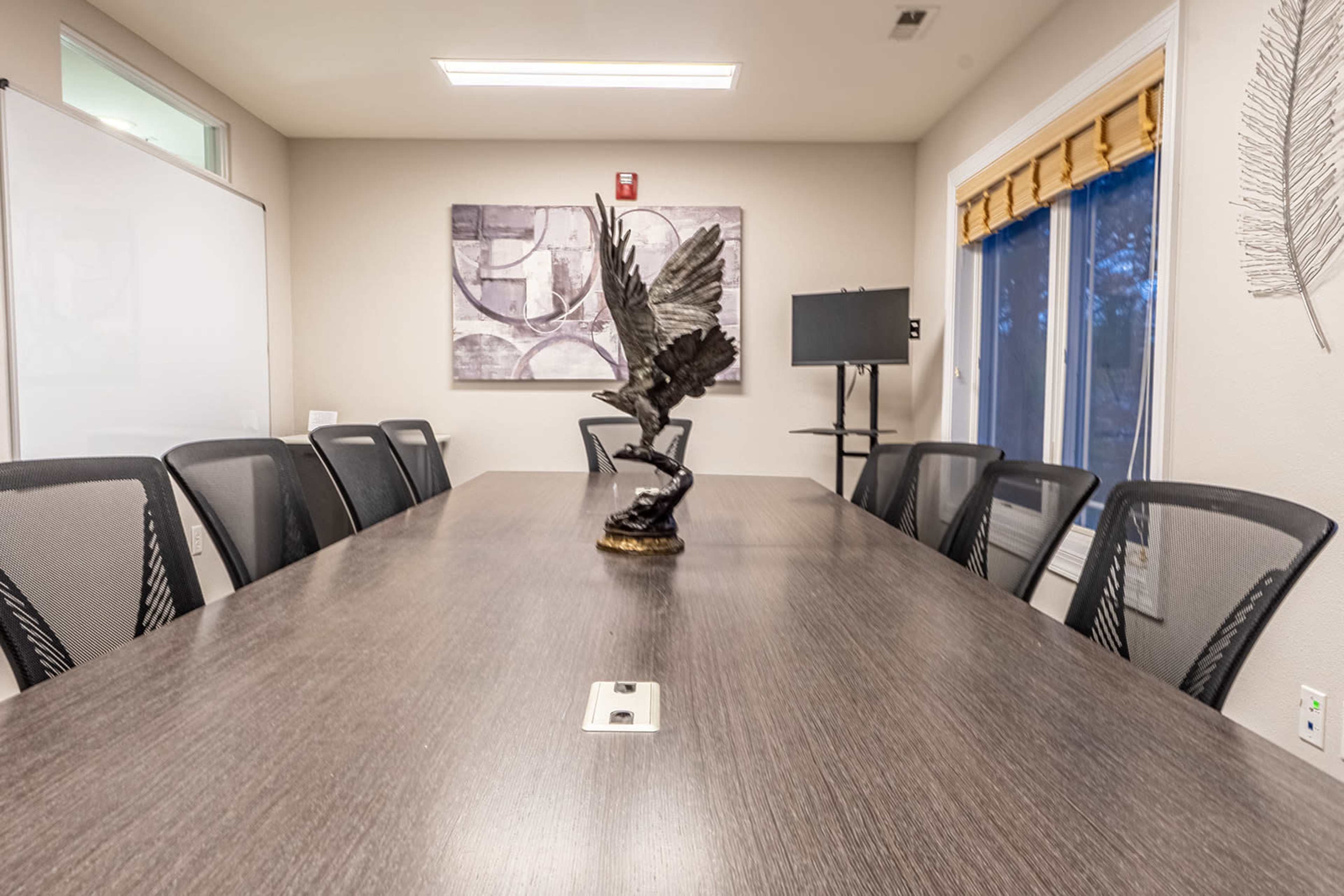 A long conference table with black chairs is set in a well-lit meeting room, featuring a decorative sculpture at its center and large windows along one wall.