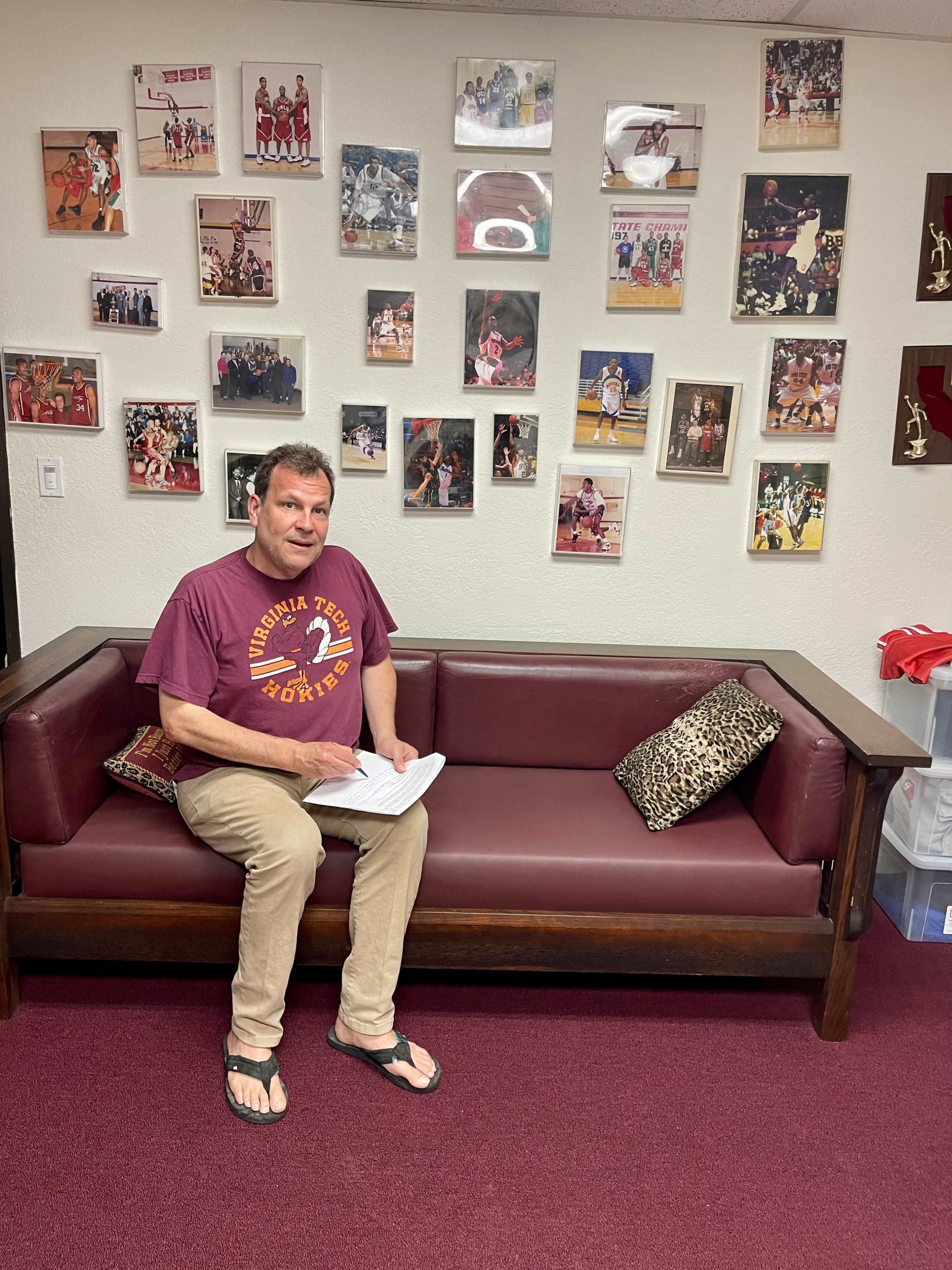A man sits on a maroon couch in a room decorated with various basketball-related photos on the walls.