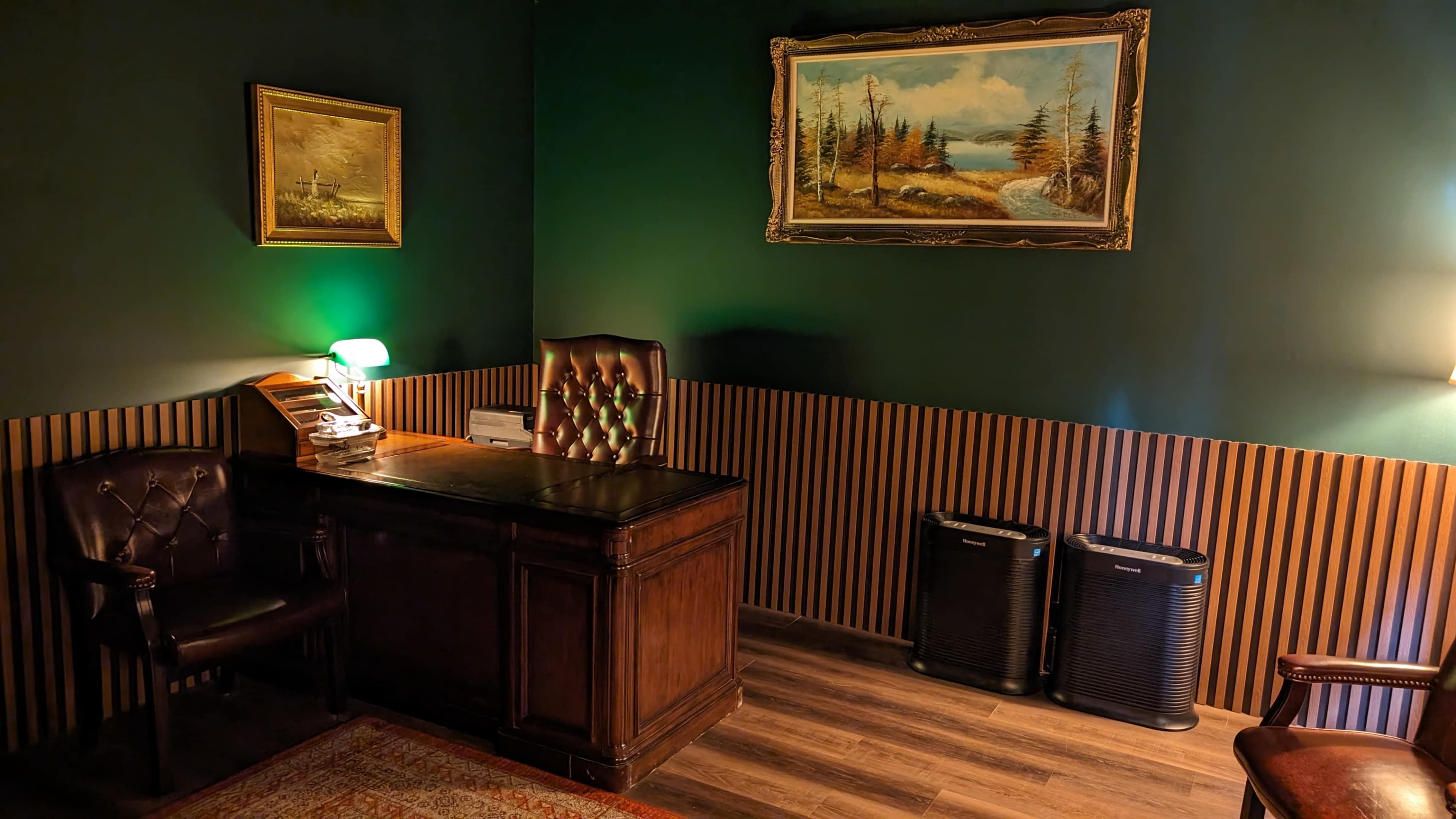 The image features a dimly lit office with a wooden desk, an upholstered chair, a painting on the wall, and two black electronic devices beside the wall.